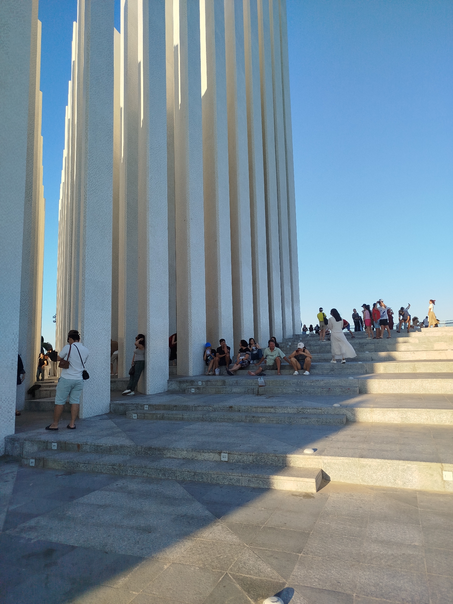 The image shows a group of people at the Landmark 81 monument in Ho Chi Minh City, Vietnam. The monument consists of tall, white, rectangular pillars arranged in parallel rows, with spaces between them. People are standing and sitting on the large, stepped platform at the base of the pillars. Some people are walking up the steps, while others are posing for pictures. The sky is clear and blue. The ground is paved with stone tiles.