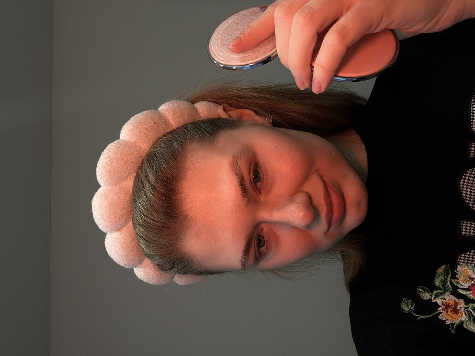A woman with a pink flower headband holds two round compacts. One of her fingers presses into the open compact's powder. She wears a black shirt decorated with a floral embroidery design and a houndstooth pattern. The background is a gray wall. The lighting casts a warm glow on the woman's face, illuminating her eyes and skin.