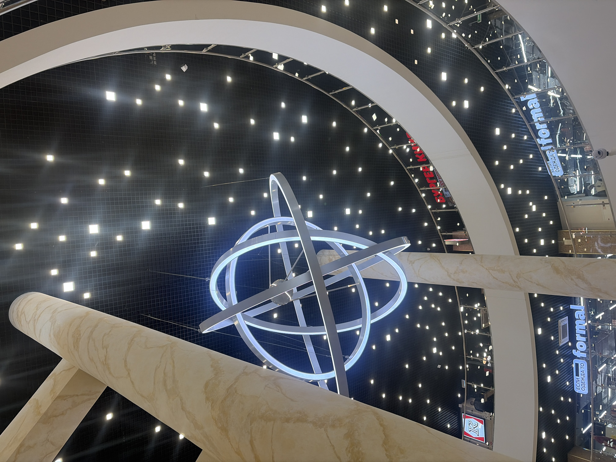 The image depicts the interior of a multi-story commercial building in Saint Petersburg, Russia, captured from a low-angle perspective looking upwards into a central circular atrium. A prominent, large chandelier, composed of multiple intersecting white rings that emit a cool blue illumination, hangs centrally from the dark, grid-patterned ceiling. This ceiling is uniformly dotted with numerous square, recessed LED lights.
Two large, cylindrical columns with a light brown, marbled finish are visible in the foreground, extending vertically through the structure. Multiple curved levels or balconies encircle the atrium, each featuring a white fascia and glass railings topped with silver handrails.
On the lowest visible level, an illuminated sign reads "ЕСЛИ ОДЕЖДА ТО formal" in white text, identifying a clothing store. Above this, a partially visible red sign with Cyrillic text is present, and on a higher level, a red square logo with a stylized white "R" is visible. Several individuals are sparsely distributed on the various levels, partially obscured by the railings, engaged in unidentifiable actions typical of a commercial environment. The overall scene conveys a modern, brightly lit, and spacious interior design.