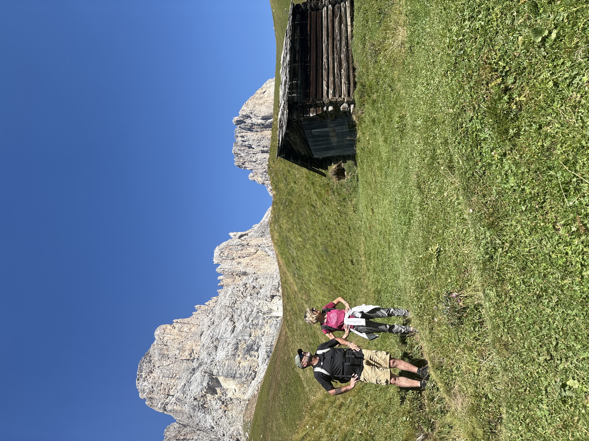 The image depicts a scenic mountain landscape in Campitello di Fassa, Italy, under a clear blue sky. In the foreground, a man and a woman are standing on a grassy slope, both appearing to be on a hike. The man, wearing a black t-shirt, tan shorts, a backpack, and a green cap, has his hands on his hips and is looking to the left. The woman, in a pink t-shirt, grey pants, and with a white jacket tied around her waist, is also standing with her hands on her hips and looking to the left. To the right of the couple and further up the slope is a rustic wooden cabin. In the background, majestic rocky mountain peaks rise sharply against the sky. The terrain is characterized by rolling green hills covered in grass and scattered vegetation. The overall impression is one of outdoor recreation and natural beauty.