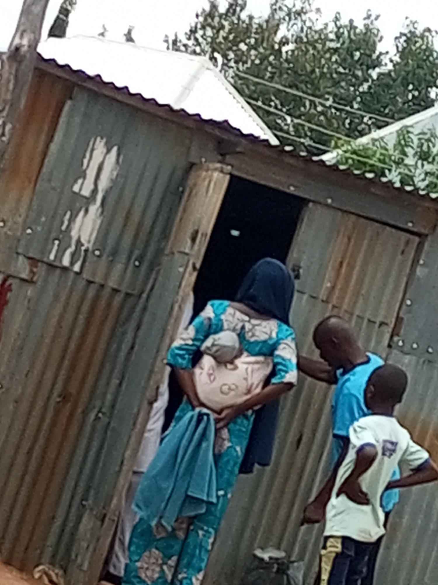 In the city of Tempure, Nigeria, a scene unfolds outside a corrugated metal structure with a white, wavy roof. Three individuals are present. A woman, facing away from the camera, wears a blue headscarf and a patterned blue and white dress, carrying a baby wrapped in a tan cloth on her back. Beside her, a man in a bright blue t-shirt has his left arm extended, possibly touching the woman or the baby. To the right, a young boy in a white t-shirt and dark pants with yellow stripes stands with his hands on his hips, looking towards the scene. A turquoise cloth is draped over the woman's shoulder and down her side. The corrugated metal walls of the structure show signs of age and weathering. In the background, green foliage is visible, suggesting a somewhat rural or less developed area.