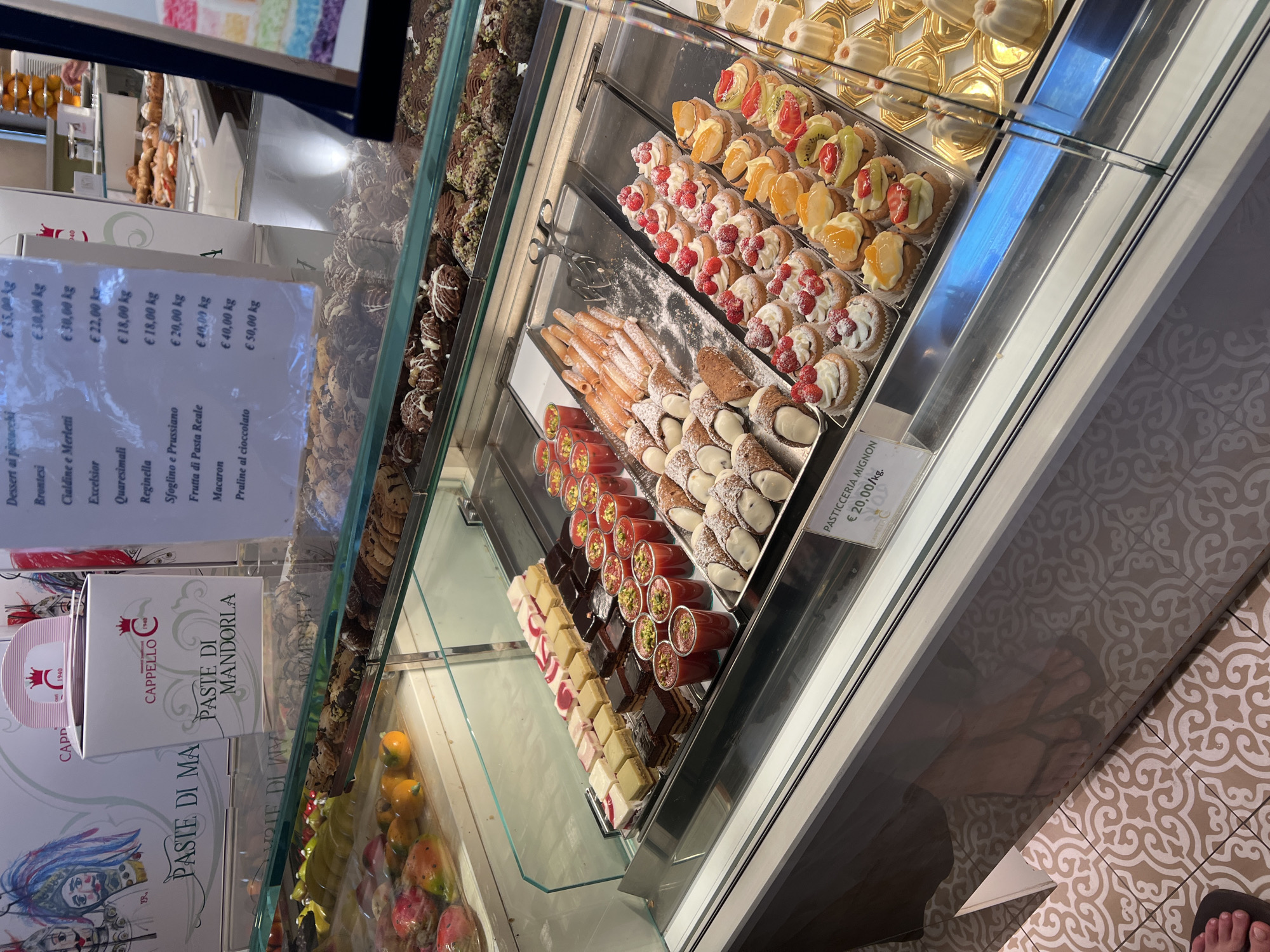 This is a photograph taken at a pastry shop in Palermo, Italy. The main subjects are the display cases filled with a variety of pastries, candies, and cookies. Visible objects include trays of cannoli, fruit-topped mini cakes, small squares of chocolate and cream desserts, and boxes of almond paste. The scene is indoors, with bright, artificial lighting illuminating the colorful and meticulously arranged treats. The overall setting is a commercial establishment, likely a "pasticceria," or pastry shop, and the overall mood is inviting and appetizing, emphasizing the abundance and craftsmanship of the baked goods.
There are no people prominently featured in the image, although a reflection of someone's feet wearing flip-flops is visible on the floor tiles near the bottom right, suggesting a customer or staff member. The time of day is not explicitly indicated but the bright lighting suggests it's likely during business hours.
Several pieces of visible text provide context:
- A price list with items like "Dessert ai pistacchi," "Brontesi," "Cialdine e Merletti," and "Macaron" listed with prices per kilogram in Euros.
- A box with the branding "CAPPELLO" and "PASTE DI MANDORLA," indicating a type of almond pastry.
- A label that reads "PASTICCERIA MIGNON" with a price of "€ 20,00/kg," identifying a selection of small pastries.
The image captures the visual appeal of Italian pastries, showcasing a rich array of traditional and modern confections. The ornate tiled floor in the foreground adds to the cultural context of the location.