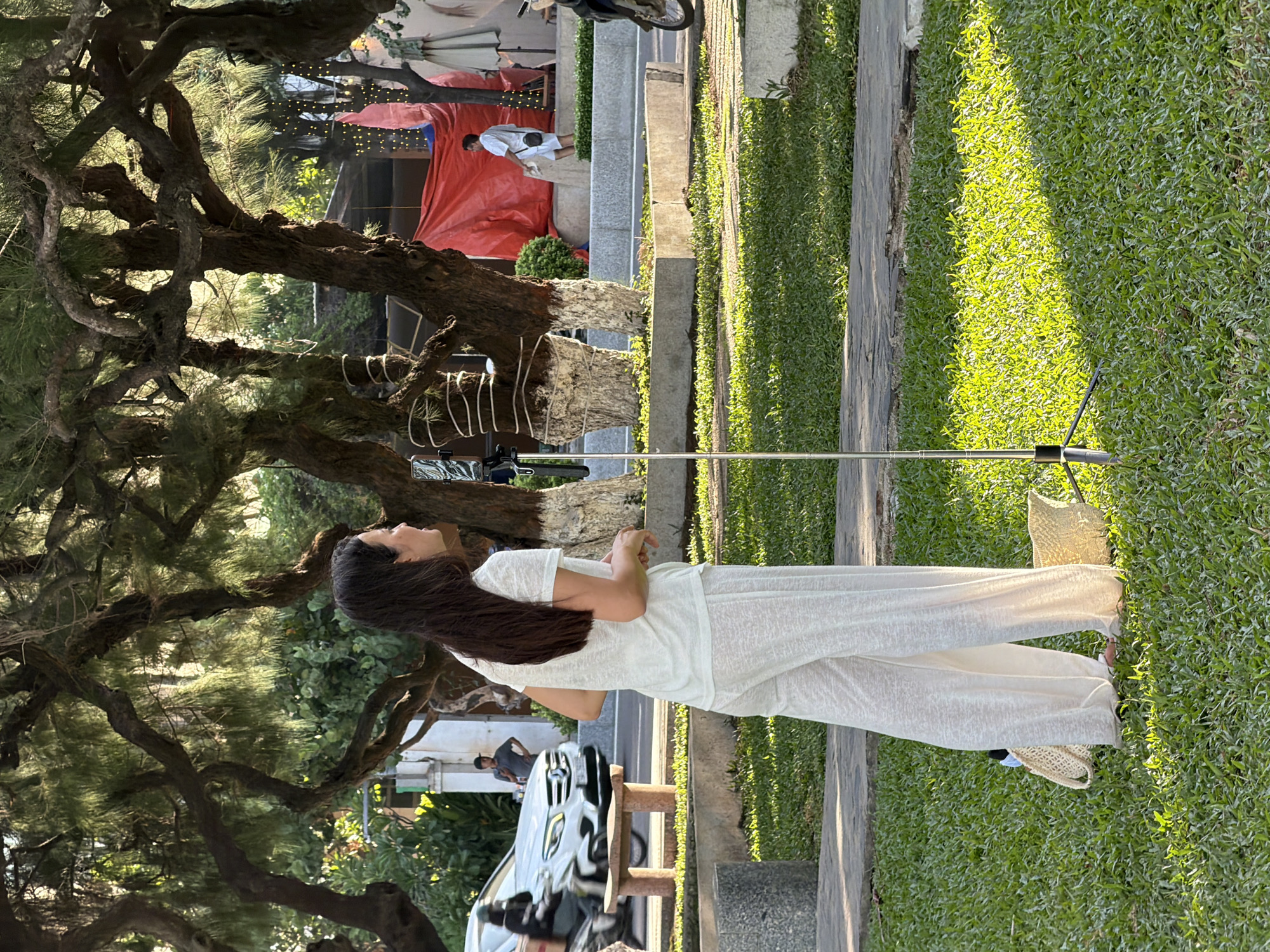 In Nha Trang, Vietnam, a young woman with dark hair, wearing a white t-shirt and loose white pants, stands on a grassy area. She is facing right, looking away from the camera, and appears to be speaking into a microphone attached to a silver stand in front of her. Two straw bags are on the grass near her feet. The scene is set outdoors, with large trees providing shade and sunlight dappling the grass. In the background, a white car is visible on a road, along with other people and structures, including a red tarp. The overall setting suggests a public park or a waterfront area.