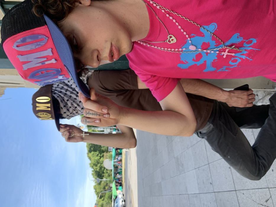 Two young people stand in front of a building. The person on the left wears a brown cap with gold letters, a black and white keffiyeh, and a brown shirt. The person on the right wears a pink shirt with a blue design, multiple necklaces, and a cap with a blue brim and pink top. Both have tattoos on their fingers. The photo is rotated to the side. The blue sky and green trees can be seen in the background.