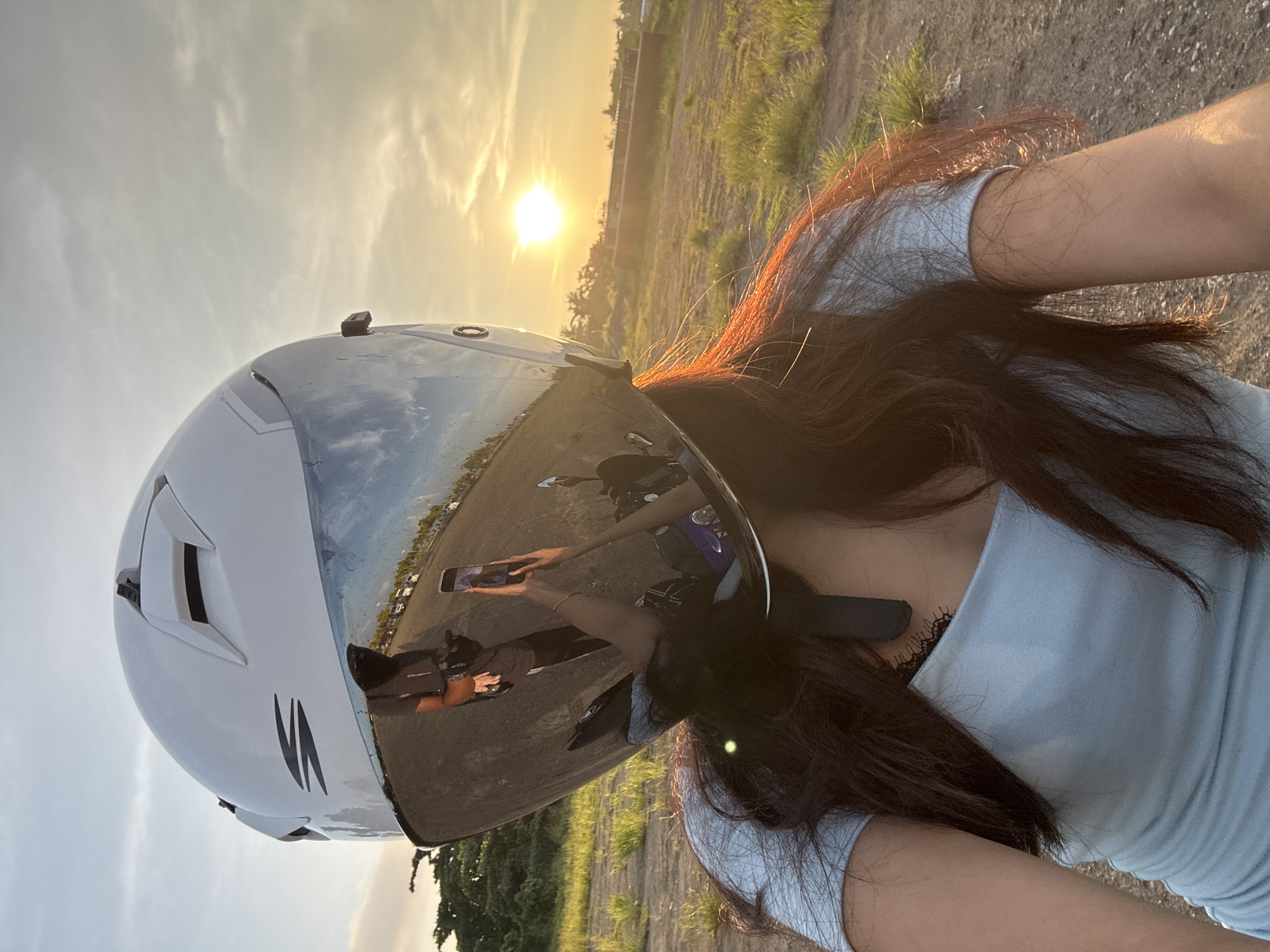 A person wearing a white helmet with a reflective visor is taking a selfie in Davao City, Philippines. The sun is setting in the background, casting a warm glow on the scene. The helmet's visor reflects the person's arm holding a smartphone, as well as a road with vehicles parked on the side and some trees and buildings in the distance. The person is wearing a light blue top. The environment appears to be an open, outdoor area with dry grass and dirt ground. The sky is clear with a few wispy clouds.