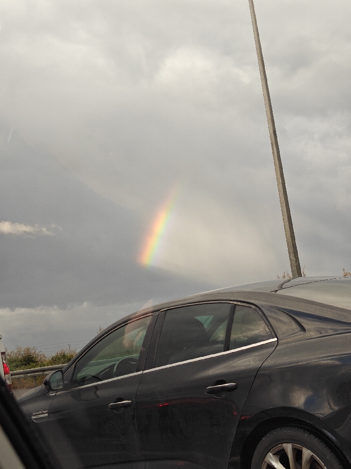 The image depicts a roadside scene in Acre, Israel, under an overcast sky dominated by gray clouds. A distinct segment of a rainbow, displaying its full color spectrum, is visible in the upper left-center against a darker cloud mass. In the midground, a dark-colored sedan, consistent with a Renault Megane Grand Coupe model, is prominently positioned. The vehicle's driver's side is visible, exhibiting slight dust accumulation on its body panels. A chrome trim piece runs along the base of the windows. A human figure, presumed to be the driver, is faintly discernible through the vehicle's front window, positioned at the steering wheel. A tall, light-colored pole, possibly a street light or utility pole, ascends vertically from the lower right towards the upper right corner of the frame. The image is captured from inside another vehicle, with a portion of the originating vehicle's interior visible in the extreme bottom left. Distant vegetation and part of another vehicle are faintly visible in the far left background.