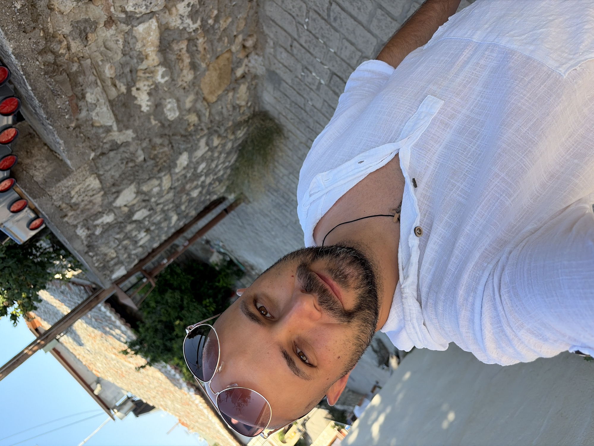A man with a shaved head, brown eyes, and a beard is taking a selfie outdoors. He is wearing a white, collared, short-sleeved shirt with buttons on the placket. He has sunglasses on top of his head, and a necklace with a pendant is visible around his neck. The background shows a stone wall and a cobblestone path, indicative of a Mediterranean setting, possibly in Kassandra Municipal Unit, Greece, given the context. The lighting suggests it is daytime.