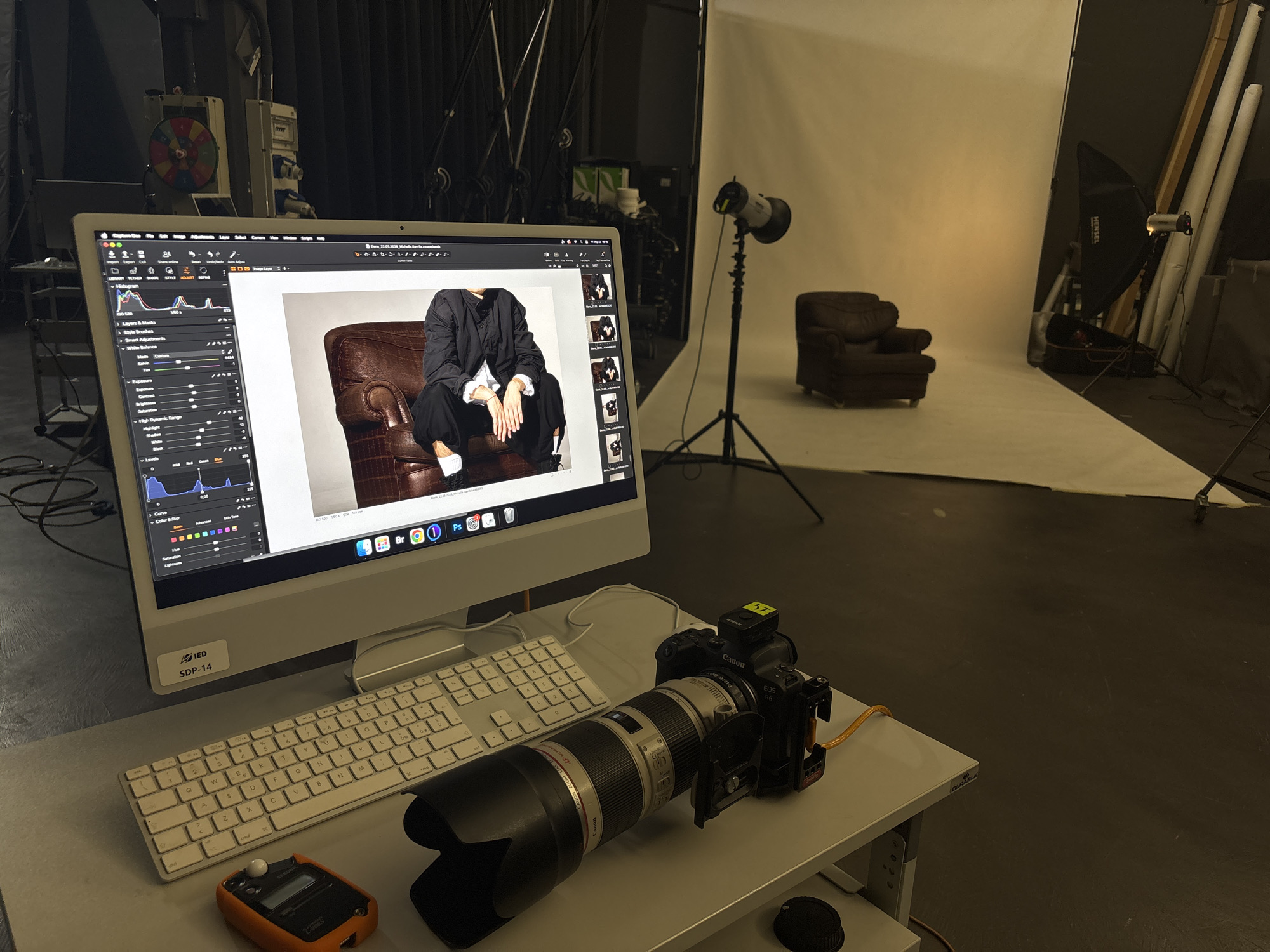 Here's a description of the image:
The image depicts a photography studio, likely used for fashion or portrait shoots. In the foreground, a white iMac computer sits on a white table, with a keyboard and a Canon EOS R6 camera equipped with a long telephoto lens resting in front. The computer screen displays image editing software with a photograph of a person seated in a leather armchair, wearing a black outfit with white socks and shoes.
In the background, a white backdrop is illuminated by studio lighting. A leather armchair sits on the white floor. Other lighting equipment, including a flash head and softbox, are positioned around the set. Various photography equipment and accessories are visible in the periphery.
Based on the "IED SDP-14" sticker on the iMac, the location is likely the Istituto Europeo di Design (IED) in Milan, Italy.