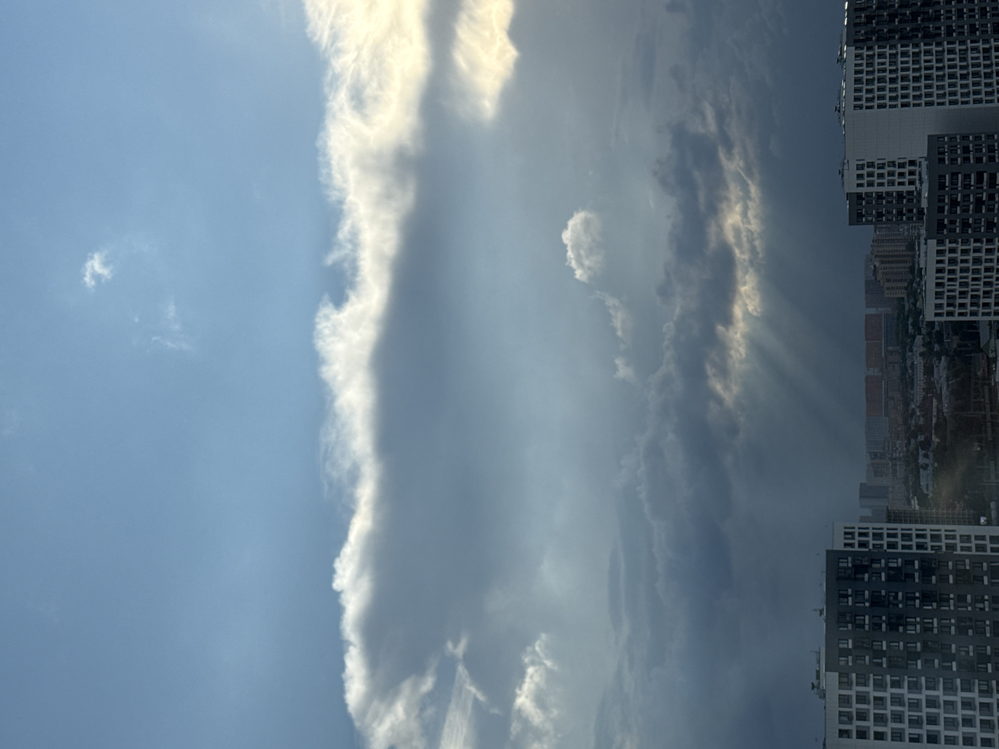 The image captures a dramatic sky over the city of Lyubertsy, Russia, as seen from an elevated viewpoint, likely a high-rise apartment building. The sky is a canvas of changing weather, with a bright blue expanse at the top giving way to large, dense clouds. A striking band of clouds, illuminated from behind by the sun, stretches across the mid-section of the frame, casting a warm, golden glow on its edges. Below this, darker, more ominous clouds gather, with visible shafts of sunlight breaking through, creating a crepuscular effect.
In the foreground, the tops of several modern, multi-story apartment buildings are visible. These buildings feature geometric patterns of windows and balconies, rendered in shades of white, gray, and dark gray. The architecture suggests a contemporary urban development. In the distance, a cityscape of more buildings, some colored in muted tones of red and yellow, stretches towards the horizon, partially obscured by the atmospheric conditions. The overall scene depicts a transitional moment, possibly before or after a storm, characterized by dynamic cloud formations and the interplay of light and shadow over an urban landscape. No people are visible in the frame.