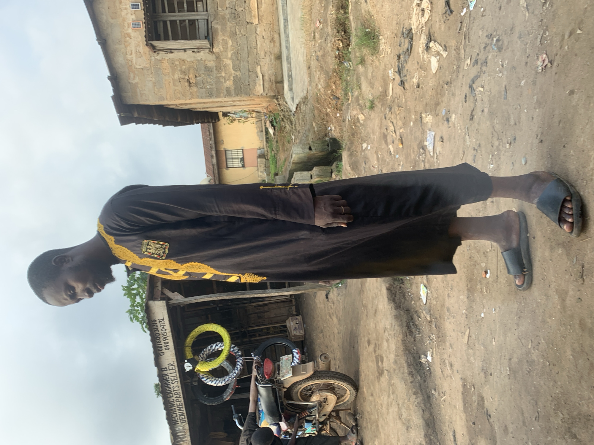 The image depicts a man standing in a dusty outdoor setting. He is wearing a long black garment with yellow embroidery on the collar and sleeves. The man is facing to the right, with his head slightly bowed. The environment appears to be a street or marketplace, with a stall visible behind him displaying motorcycle tires. A motorcycle is also parked nearby. In the background, there are buildings with weathered facades. The ground is uneven and covered with dirt and debris. The sky is overcast. The scene is likely in Shagamu, Nigeria, as indicated by the context provided.