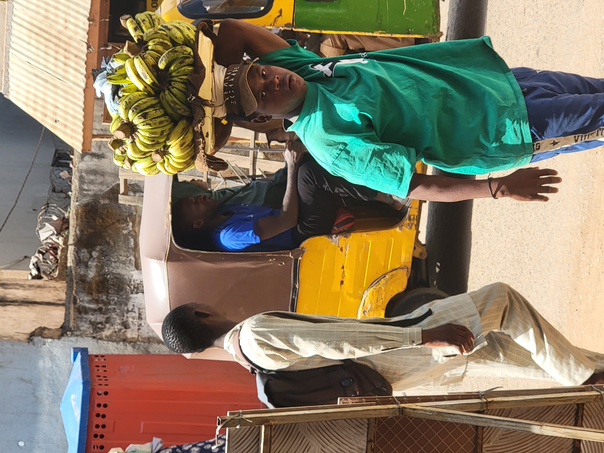 The image captures a vibrant street scene in Maiburiji, Nigeria, under bright daylight. The central figure is a young person, likely a vendor, carrying a large, impressive bunch of green bananas balanced expertly on their head. They are wearing a faded green t-shirt with a white graphic, a light brown cap, and blue track pants with "VUITTO" visible on the side, likely part of a "LOUIS VUITTON" branding. This person looks directly at the camera with a neutral expression.
Behind and to the left of the main subject, another person is seated inside a yellow and green auto-rickshaw, commonly known as a keke Napep in Nigeria. This individual is wearing a blue shirt. In the foreground, partially obscuring the auto-rickshaw, stands a third person seen from behind, wearing a light-colored, possibly striped, long-sleeved shirt and trousers, with a dark backpack.
The background shows various elements of an urban or semi-urban environment. A large red container or structure is visible on the left, along with what appears to be part of a building with a corrugated metal roof. The ground is dusty, indicating a typical street setting. The strong sunlight casts clear shadows, suggesting mid-morning or afternoon. The overall impression is one of daily life and commerce in a bustling locale.