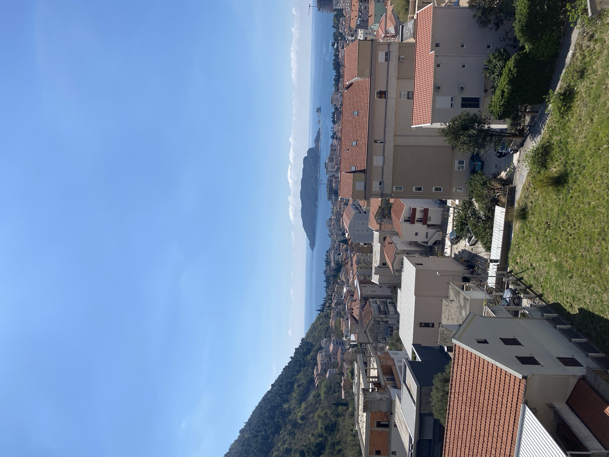 A high-angle, outdoor photograph reveals a cityscape and coastal view in Budva, Montenegro. The scene, lit by daylight, has a clear, blue sky. Buildings, predominantly with red-tiled roofs, occupy the lower right and midground, extending toward the coastline. Vegetation covers a hillside to the lower left. The Adriatic Sea is visible in the distance, with a landmass, likely the island of Sveti Nikola, positioned in the center of the view.