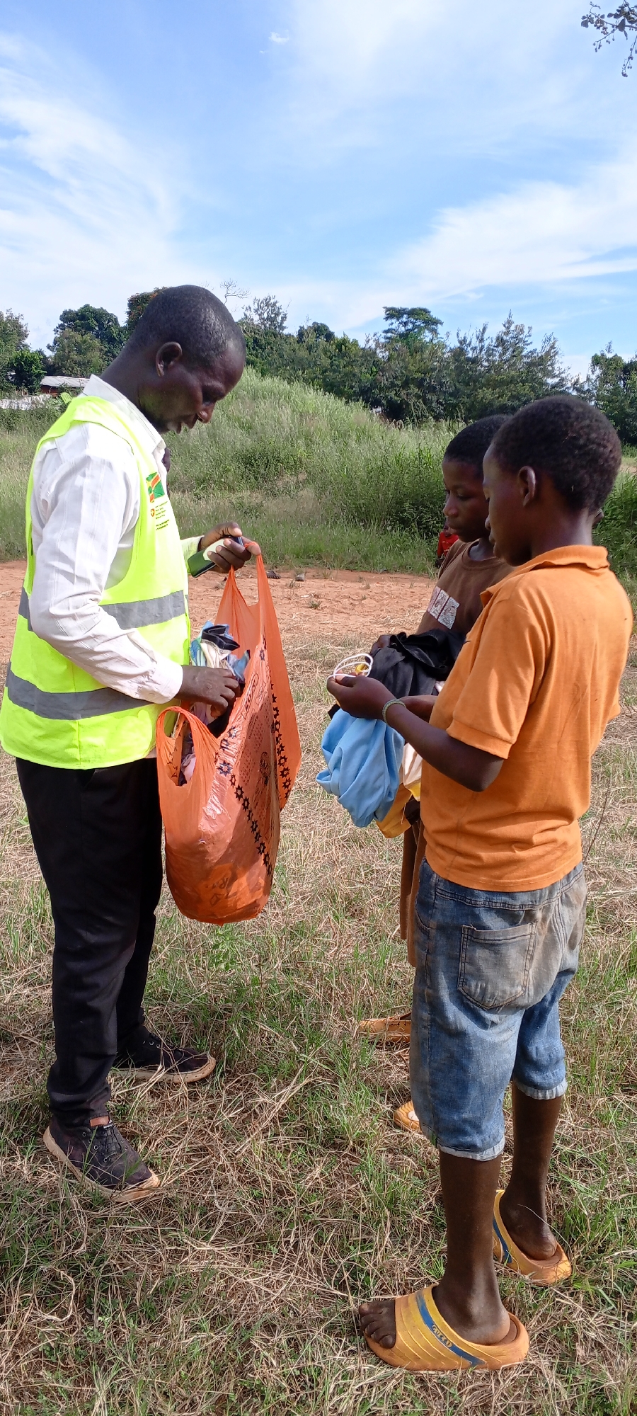 In Ngalula, Democratic Republic of the Congo, an adult male wearing black pants, black shoes, a white shirt, and a yellow safety vest, is holding an orange plastic bag filled with clothing in a grassy field. Two children stand opposite the adult, one wearing an orange shirt and jeans and the other a brown shirt. One child is holding a piece of blue cloth. The background includes green trees, bushes, and a blue sky with clouds.