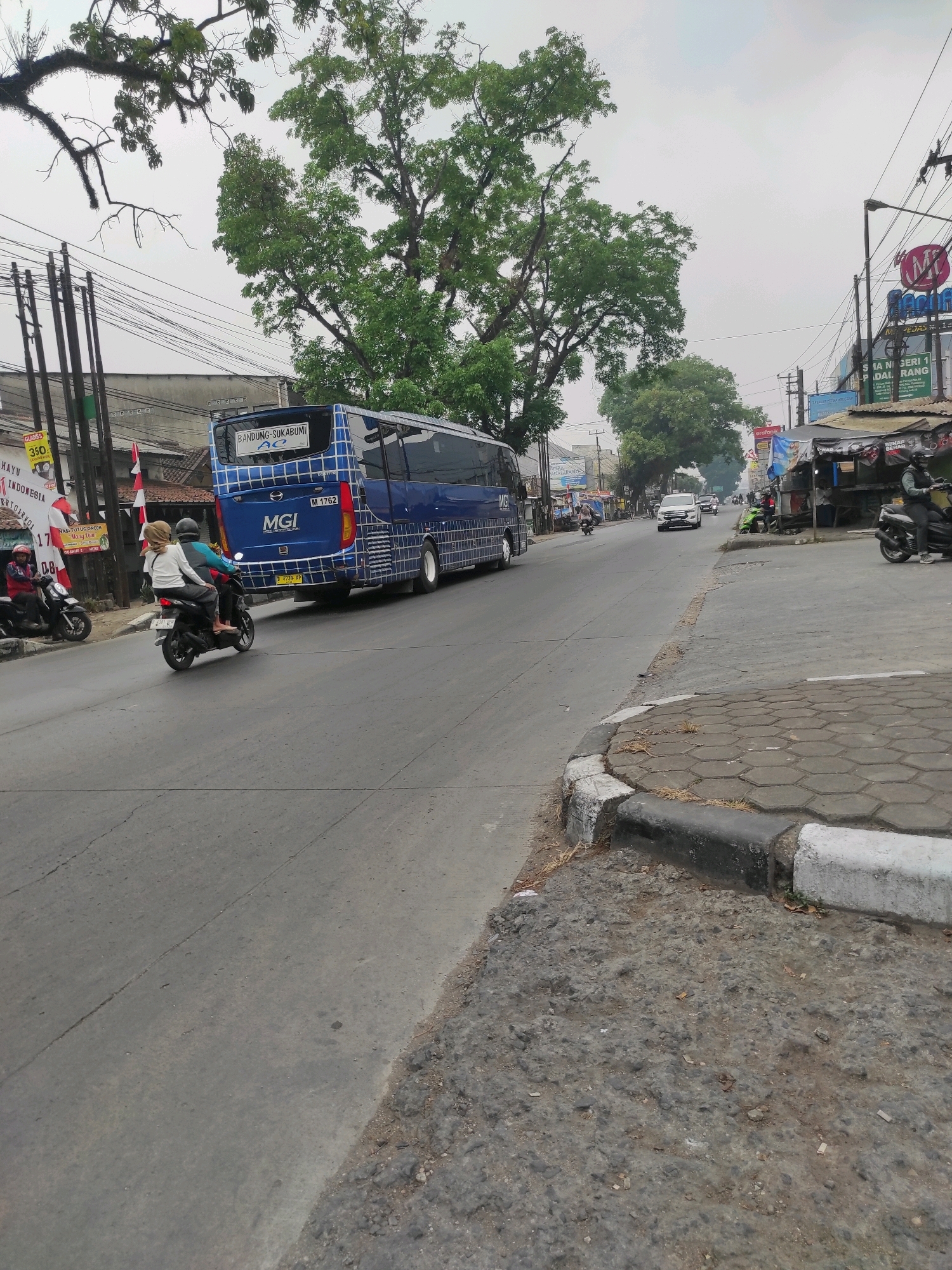 The image captures a street scene in Padalarang, Indonesia, on an overcast day. A blue MGI bus with "BANDUNG-SUKABUMI" written on its back is the dominant subject, traveling away from the viewer. In front of the bus, two individuals are riding a motorcycle, with the passenger wearing a light-colored hijab. To the left of the bus, a motorcycle is parked on the side of the road, with a person wearing a red jacket seated on it. The road appears to be paved and shows signs of wear. On the right side of the road, there are various small shops and stalls, some with awnings and signs. Power lines are visible overhead, supported by utility poles. Lush green trees are scattered throughout the scene, particularly a large tree to the left of the bus and smaller trees in the distance. The overall environment suggests a bustling street with active transportation.
