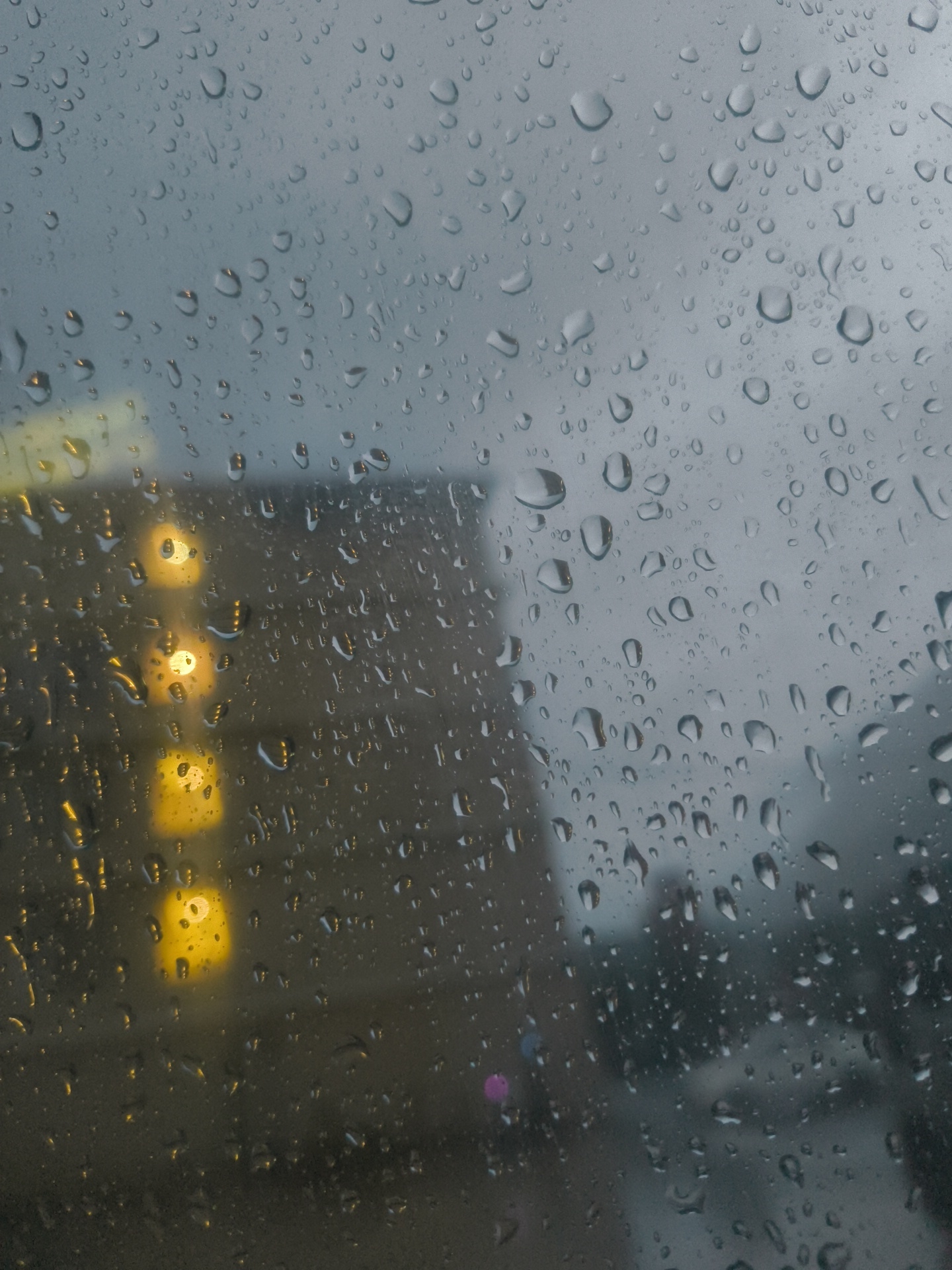 The image depicts a view through a rain-streaked window, likely from a vehicle or building. Large, rounded raindrops cling to the glass, distorting the scene beyond. In the background, a building with several illuminated windows is visible. The windows appear to be a warm, yellow-orange hue. The overall environment suggests a dreary, possibly urban setting, with the building implying a location within a city. The rain creates a sense of introspection and isolation.
