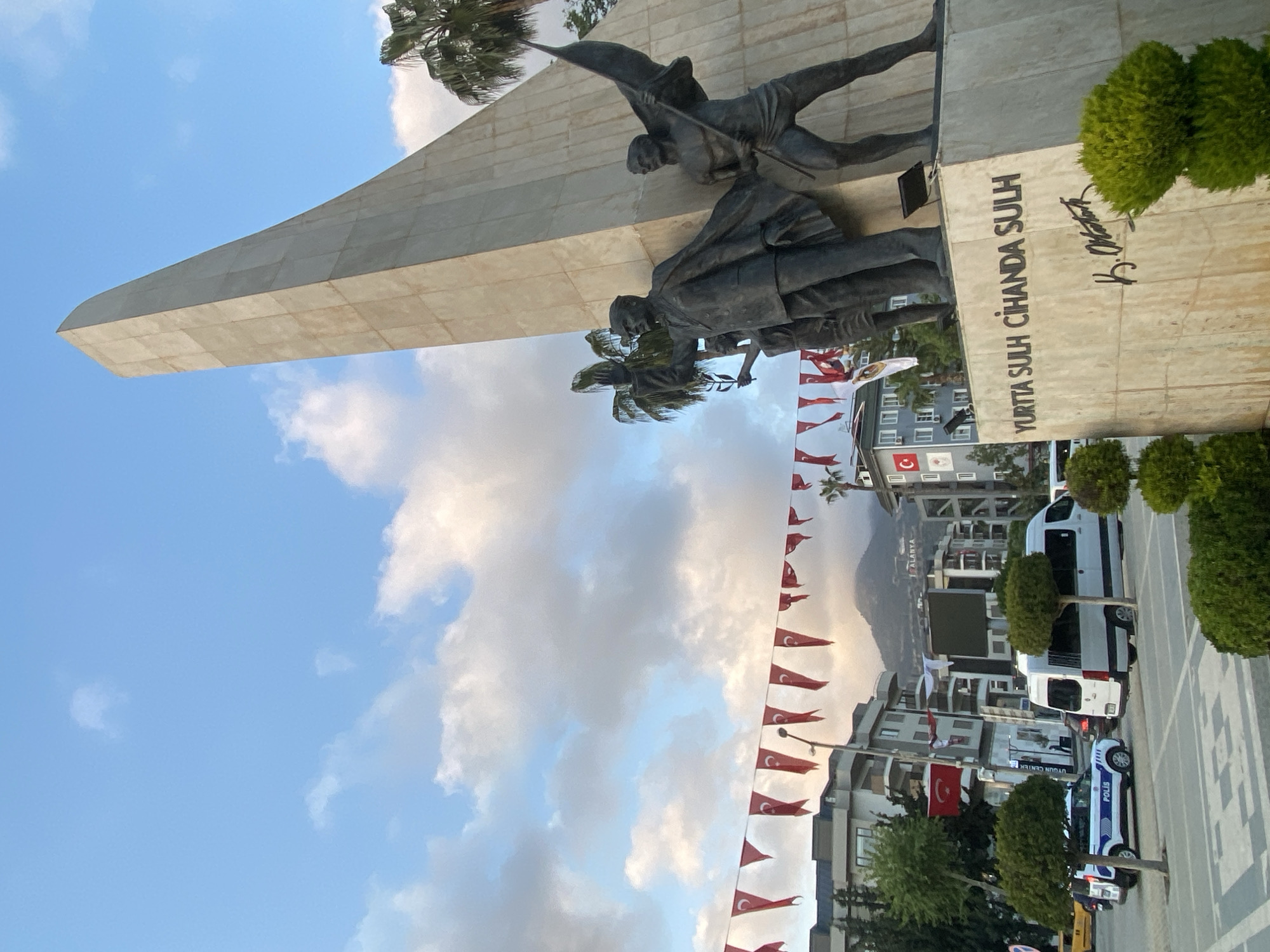The image, when viewed upright, displays a grand monument in a public space in Alanya, Turkey, under a clear blue sky with scattered clouds.
**Subjects:** The central feature is a large, angular stone monument, likely made of concrete or light-colored stone. Adorning the monument are several bronze statues. One prominent figure is depicted dynamically, seemingly moving forward while holding a flag-like object aloft. Below this figure, another statue portrays a person in a seated or kneeling posture. Further figures are partially visible within the sculptural group. Lush palm trees grow near the monument. In the surrounding urban environment, there are multi-story buildings, green shrubbery, and parked vehicles.
**Scene and Setting:** The monument stands in what appears to be a town square or a wide street. The background reveals urban architecture, including modern buildings with visible balconies. In the mid-ground, several vehicles are parked, including white vans and a white police car marked "POLİS." Numerous small Turkish flags, red with a white star and crescent, are strung across the street, adding a festive or nationalistic touch. A larger Turkish flag flies from a building. Distant mountains frame the horizon, with the word "ALANYA" visibly spelled out on one of the slopes, likely a large sign.
**Activity or event taking place:** There is no specific activity or event being depicted; the scene captures a static view of a public landmark. The arrangement of flags suggests a general celebratory atmosphere or national pride.
**Notable details:** The time of day appears to be daytime, with bright, natural lighting. The sky is largely blue with some patches of white and grey clouds. The presence of the "POLİS" vehicle, the Turkish flags, and the "ALANYA" sign on the mountain clearly indicate the location as Alanya, Turkey.
**Visible text:** Engraved on the base of the monument is the Turkish phrase "YURTTA SULH CİHANDA SULH," which translates to "Peace at Home, Peace in the World." Below this, there is a distinct signature, identifiable as that of K. Atatürk (Mustafa Kemal Atatürk). The name "ALANYA" is also visible on the distant mountainside.