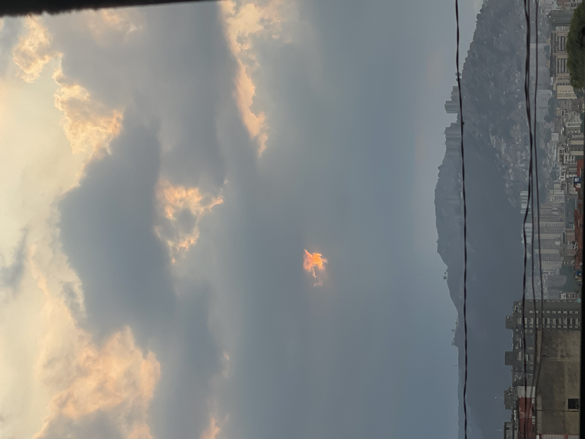 The image shows a view from a window in Caracas, Venezuela. The sky is filled with gray clouds, with some clouds tinted orange by the setting sun. In the center of the sky is a small, bright orange cloud that resembles a fire. To the right of the clouds is a mountain range, partially obscured by haze. In front of the mountain range are buildings and power lines. The bottom right corner of the image shows a part of the window frame.