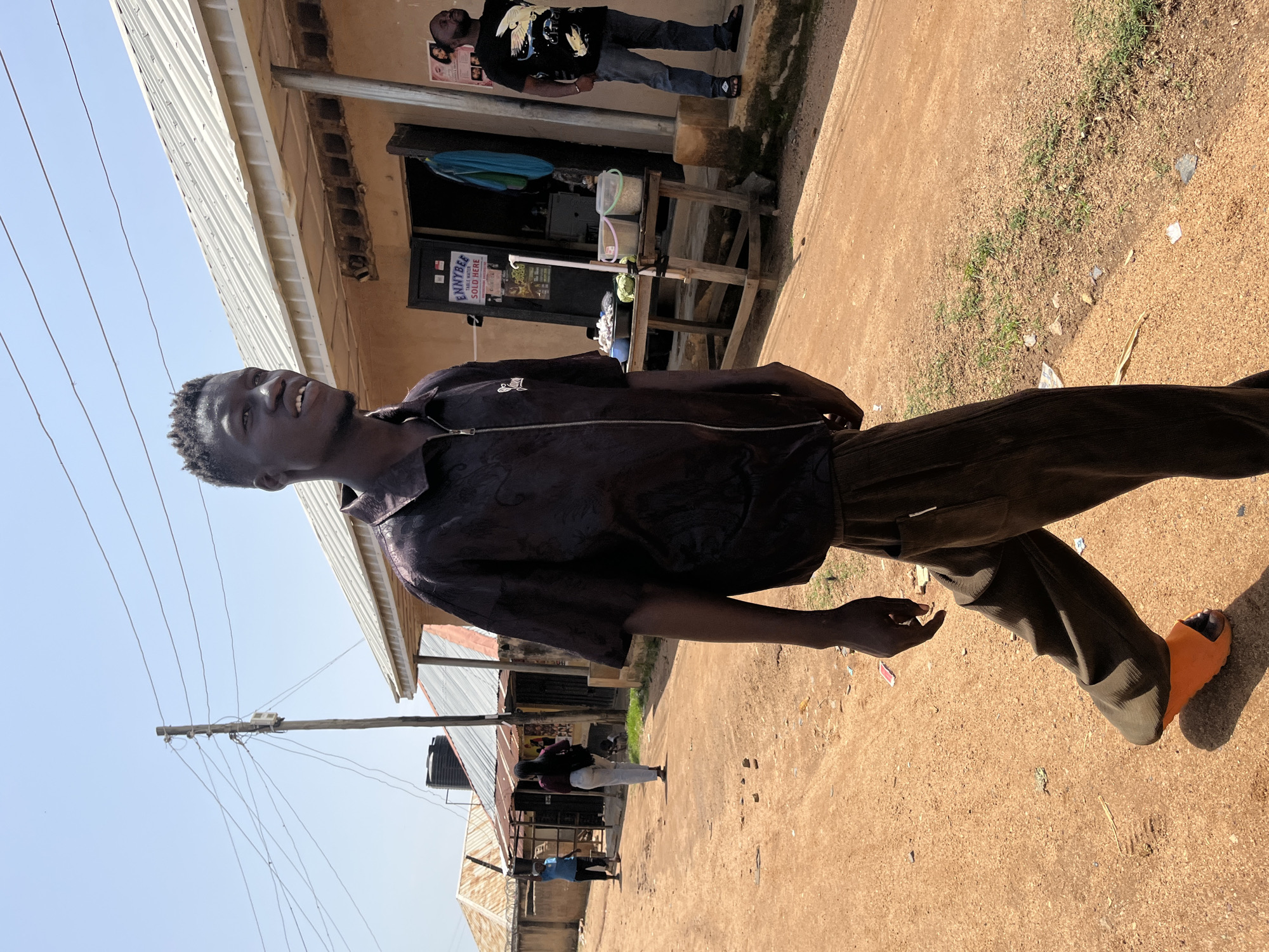 A young Black man is walking on a dirt road in Jos, Nigeria, with a store and other buildings in the background. He is wearing a dark, short-sleeved, collared shirt with a zipper and dark, loose-fitting pants. His right foot is adorned with an orange slide sandal, while his left foot is not visible. The man has a determined expression as he looks to his right, mid-stride. To the left of the man, another man in a black t-shirt and blue jeans stands near the store's entrance. Further down the road, two other individuals are walking away from the camera. The store's exterior features a metal roof, a wooden table with various containers, and several posters on the wall. The ground is covered in reddish-brown dirt, with sparse patches of green grass and scattered debris. A wooden utility pole with electrical wires stands tall against the clear, bright sky, suggesting a sunny day.