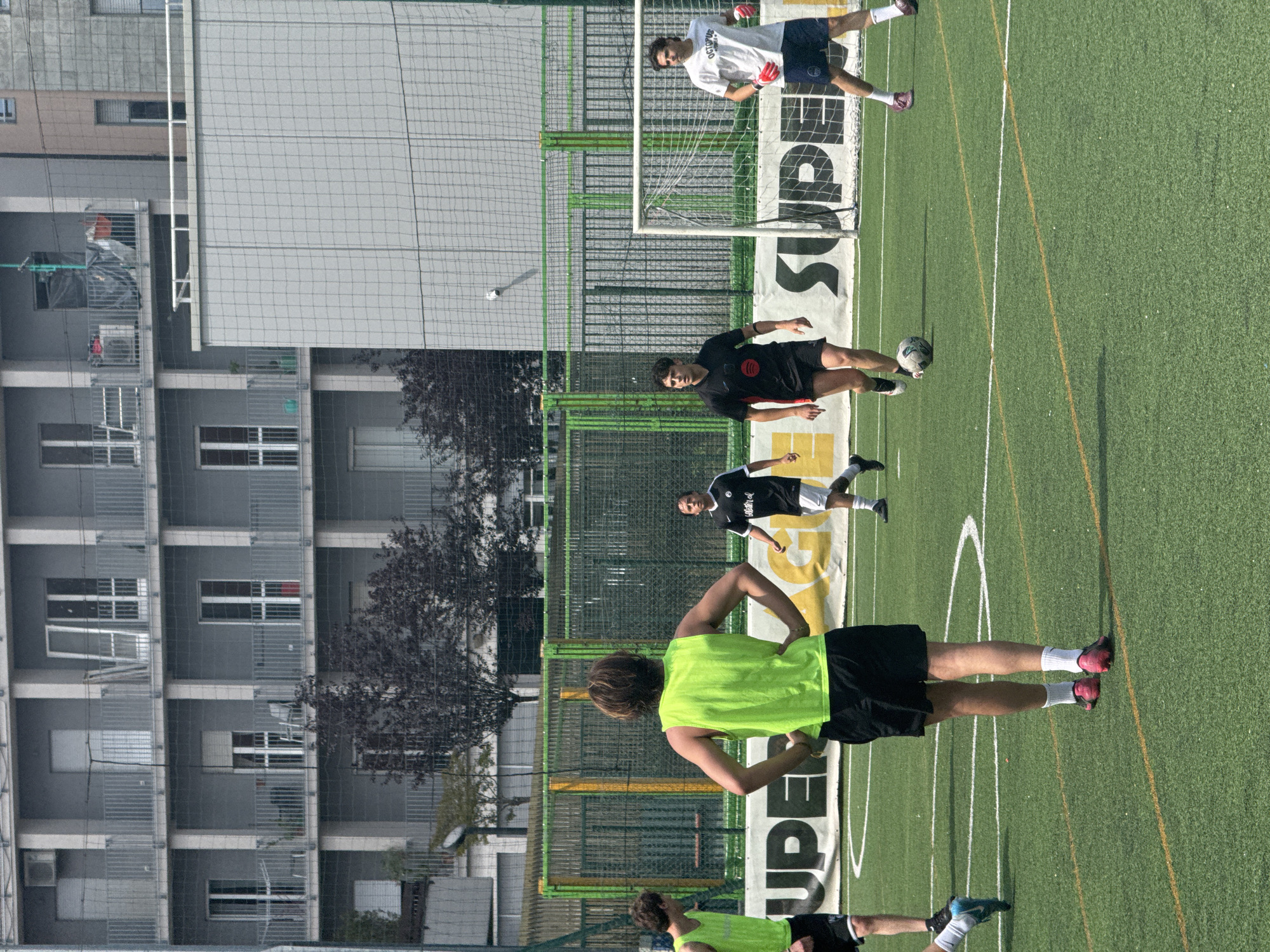 An outdoor artificial turf football pitch in the city of Milan, Italy, is observed under clear, sunny conditions. Five males are visible on the green playing surface. In the foreground, a male with brown hair, wearing a neon green tank top and black shorts with pink football boots, stands with his back to the camera, hands on hips. To his left, a second male, partially cropped, wears a similar neon green tank top and black shorts with blue and white football boots. In the mid-ground, a male wearing a black short-sleeved jersey with red accents and a white logo, along with black shorts and boots, stands near a white and black football. To his right, a fourth male wears a black and white striped short-sleeved jersey with a white logo, white shorts, and black boots. On the far right, positioned near a white metal football goal with a net, a fifth male wears a white t-shirt with "OCTOPUS" text, dark blue shorts, and red goalkeeper gloves, wearing pink/purple boots. The pitch features white demarcation lines and a central yellow line. Behind the pitch, a white banner displaying partial black lettering "UPE" on the left and "SUPE" on the right is visible, with a yellow banner section behind it. A green metal fence and dark green protective netting separate the playing area from the background. Beyond the fence and dark-leaved trees, multi-story grey residential buildings with numerous balconies and windows are visible. One top-floor balcony features an air conditioning unit, and another displays an open door. A fine mesh net is observed covering portions of the background buildings.