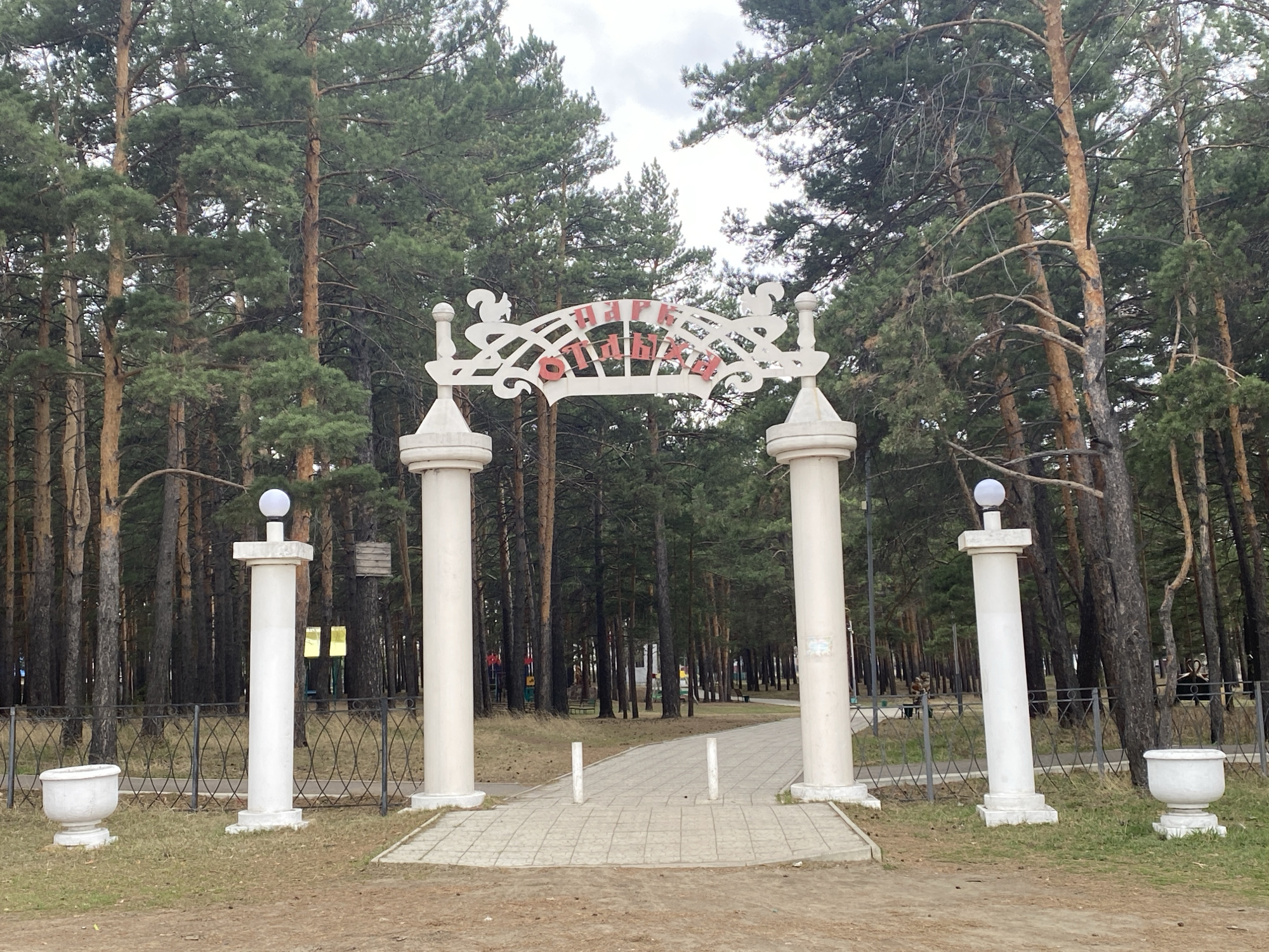 The image shows the entrance to the Park of Culture and Recreation in Sosnovka, Russia. The entrance is marked by a decorative white archway with two large columns on either side. Smaller columns with spherical lights flank the entrance on the outer edges. The archway has a decorative design with stylized squirrels and the words "Парк Отдыха" (Park of Recreation) written in Cyrillic. A paved pathway leads into the park through the archway. The park itself is heavily wooded with tall pine trees. A decorative wrought iron fence runs along the perimeter of the entrance area, and white planters sit on either side of the outermost columns. The ground is covered with a mix of grass and brown leaves. The sky is overcast. People can be seen in the background, walking on the pathway inside the park.