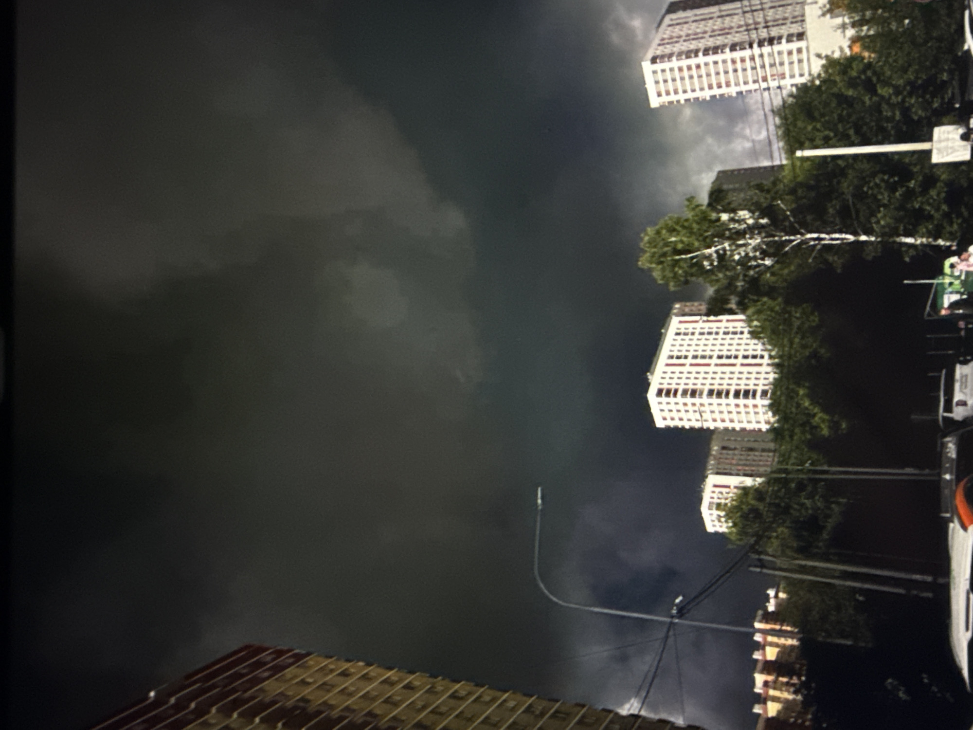 The image captures a street-level view of a city environment under a dark, ominous sky, suggesting impending bad weather. In the foreground, the roof of a white vehicle with an orange accent is visible. To the right, a white vehicle is parked. Further into the scene, several residential buildings are present. On the far left, a tall, yellow-sided building with balconies is partially visible. In the middle ground, a cluster of buildings is seen, one with a red-brown facade and balconies, and another taller white building with red vertical accents and numerous windows. A distinct, tall white birch tree stands in front of a darker building, with a streetlamp arching over the scene from the left. The sky is dominated by heavy, dark clouds, with lighter patches indicating the presence of sunlight struggling to break through. Dense green trees line the base of the buildings. The overall impression is one of a somber, overcast day in an urban setting.