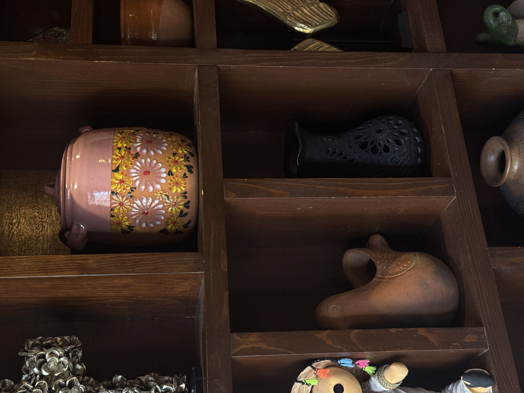 A close-up shot shows a dark wooden shelving unit containing various decorative objects. On the left side of the unit, there is a pink vase with a floral pattern. To the right, there is a black vase with cut-out patterns. Below the black vase, a brown, possibly clay, object shaped like a bird or animal is visible. In the bottom left, there's a collection of shells or small white objects. At the bottom right, portions of what appear to be handmade dolls with colorful details can be seen. The photo was taken in San Andrés Cholula, Mexico.