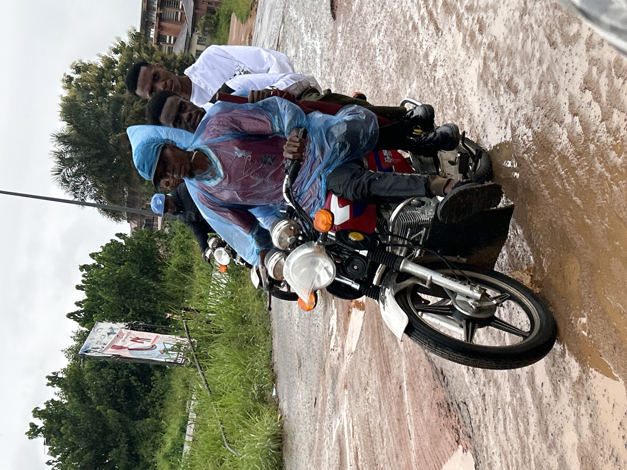 This image depicts a rural or semi-urban scene in Ihiagwa, Nigeria, featuring multiple individuals on motorcycles navigating a severely muddy, unpaved road under an overcast sky.
In the foreground, a red motorcycle carries three dark-skinned males. The driver wears a light blue disposable rain poncho over a red shirt with a visible "LFC" logo (Liverpool Football Club), black pants, and dark shoes. A blue cap is visible beneath the poncho's hood. His hands are on the motorcycle's handlebars, which feature silver grips and amber turn signals. Directly behind the driver, a second male, also dark-skinned, is seated, wearing a white shirt and dark pants. His face is partially obscured. A third dark-skinned male occupies the rear seat, wearing a white shirt, looking directly towards the camera with a serious expression. He holds a dark, vertical, stick-like object, possibly a folded umbrella or a pole, between himself and the middle passenger. The front motorcycle has a silver front fender, a clear headlight, and silver wheel spokes.
Further in the background and slightly to the left, a second motorcycle is visible, with at least two individuals. The driver, a dark-skinned male, wears a dark jacket and a blue baseball cap.
The road surface is extensively covered in reddish-brown mud and standing water, showing numerous tire tracks, indicating significant deterioration and wet conditions. Dense green foliage, including trees and tall grasses, lines both sides of the road. Two leaning billboards, partially obscured by the vegetation, are visible on the left side of the frame. In the far background, beneath the tree line, brown-colored architectural structures are discernible. A dark utility pole extends vertically on the left side of the image. The ambient lighting is flat and diffused, consistent with an overcast day.