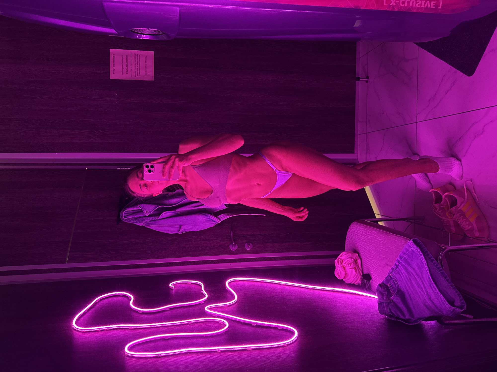 A woman in a light-colored bikini lies on a dark floor in what appears to be a tanning salon, taking a selfie with her phone. A bright pink neon light in the shape of clothing hangers lies on the floor. A denim jacket is draped nearby. To the right, a metal rack holds a pair of pink and orange sneakers and a pair of denim shorts. A sign is visible on the wall in the background. Given the Cyrillic script, the location may be Saint Petersburg, Russia.