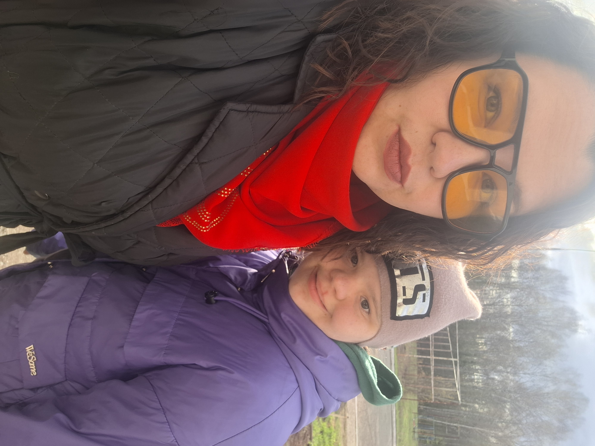 The image depicts two individuals, likely a mother and child, in an outdoor setting in Марковский, Russia. The mother is wearing a black quilted jacket, a red scarf, and large yellow-tinted glasses. The child is wearing a purple puffer jacket with a green hood and a gray beanie with a silver patch. The background shows trees and a fence, indicating a park or residential area. The lighting suggests it is daytime, with a slight glare from the sun. The image appears to be a selfie or casual snapshot.