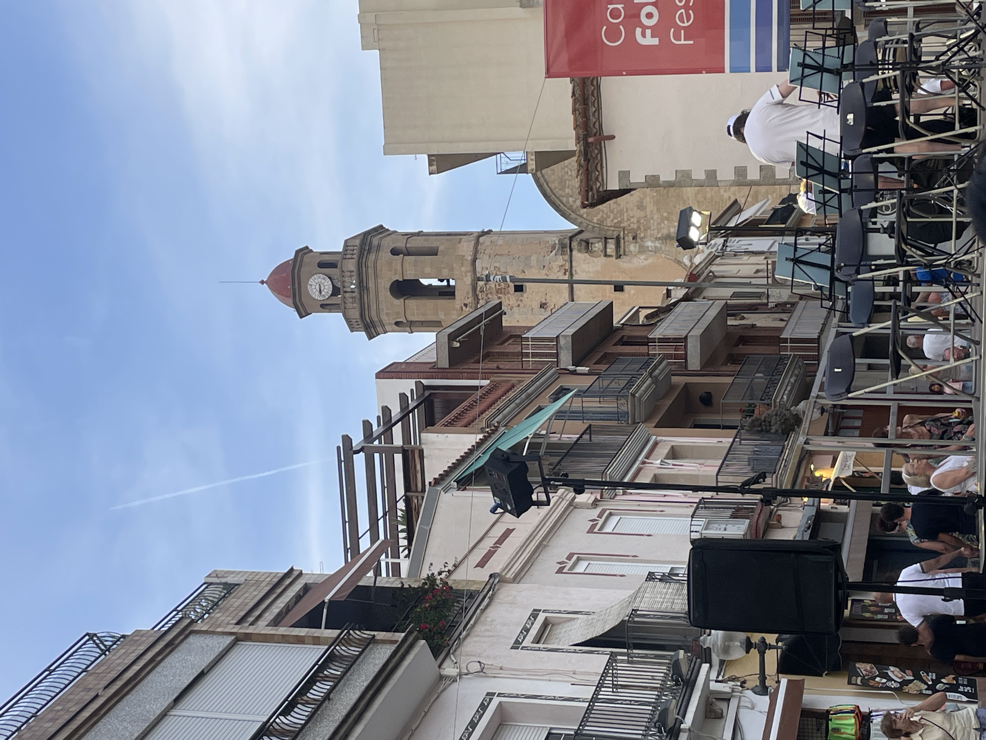 The image captures a street scene in Calella, Spain, possibly during an event or festival. In the background, a prominent clock tower with a red dome stands tall against a hazy blue sky with a faint contrail. The tower appears to be part of an old church or civic building. The foreground is occupied by a street with multi-story buildings on the left, featuring balconies with railings and awnings. On the right, a large red banner with white text, partially visible, suggests a public event.
In the middle ground and foreground, a group of people are gathered. Several individuals are standing, some looking towards the right side of the frame. A stage or performance area is set up with numerous empty chairs and music stands, implying a musical performance is about to happen or has just concluded. One person wearing a white t-shirt and a cap is seen standing behind the music stands, facing away from the camera. Other individuals are positioned in front of the stage area, some appearing to be spectators or participants. The overall atmosphere suggests a lively outdoor gathering in a Mediterranean town.