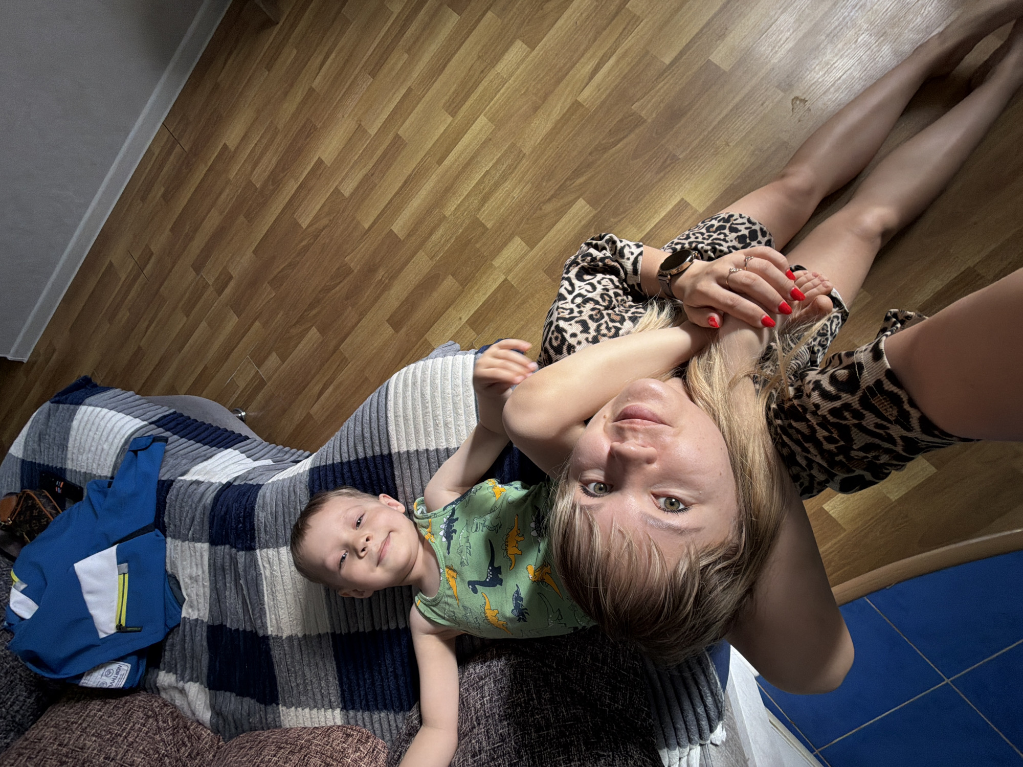 The image shows a woman and a young boy lying on a bed or couch in what appears to be an indoor setting. The woman has blonde hair and is wearing a leopard-print top. She is looking directly at the camera. The boy is wearing a green dinosaur-patterned tank top and is lying next to her. The bed is covered with a blue and grey striped blanket, and a blue jacket is placed on top. The floor in the background appears to be made of wooden planks, and part of the wall shows blue tiles. The location, Chaikovsky, Russia, cannot be independently verified based on the image alone. There is no additional context or specific details in the image to pinpoint the exact location within the city.