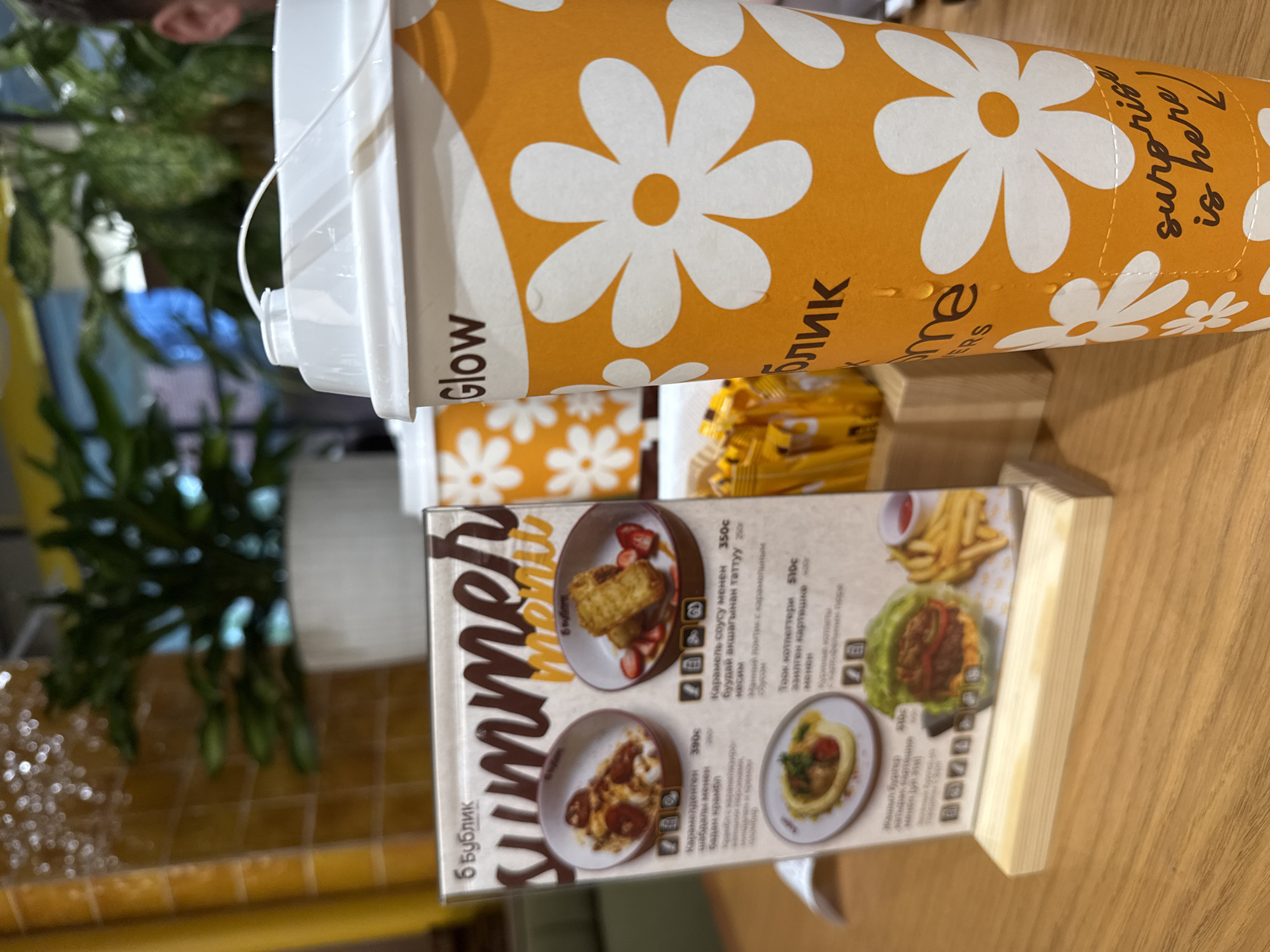 The image, rotated 90 degrees counter-clockwise, displays a close-up of a beverage cup and a menu on a wooden table, likely in a cafe or restaurant setting. In the blurred background, green foliage suggests indoor plants, and a person's ear is partially visible on the left.
The most prominent object is a tall, orange paper cup with a white plastic lid and a small handle. The cup is adorned with a pattern of white daisy-like flowers. Visible text on the cup includes "Glow" on the left side, Cyrillic script "блик" (likely part of "Бублик") in the center, and "surprise is here!" in a handwritten style on the right. Small water droplets are visible on the cup's surface.
In front of the cup is a small, freestanding menu titled "б Бублик" (b Bublik) at the top and "Summer menu" below it. The menu features several food items with accompanying pictures, descriptions in Cyrillic script, and prices. Dishes include "Карамелденген шабдалы менен Бадам крамол" (Caramelized peach with almond crumble) for 390c, "Карамель соусу менен Фуудай акшағынан таттуу кесим" (Sweet slice with caramel sauce from Fuda wheat) for 350c, "Тоок котлеттери эзилген картешке" (Chicken cutlets with mashed potatoes) for 510c, and "Жашыл Бургер" (Green Burger) also for 510c. Small icons are placed next to each menu item, possibly indicating ingredients or dietary information. Behind the cup and menu, several small, yellow packets, likely sugar or condiments, are stacked. The overall scene suggests a casual dining experience.