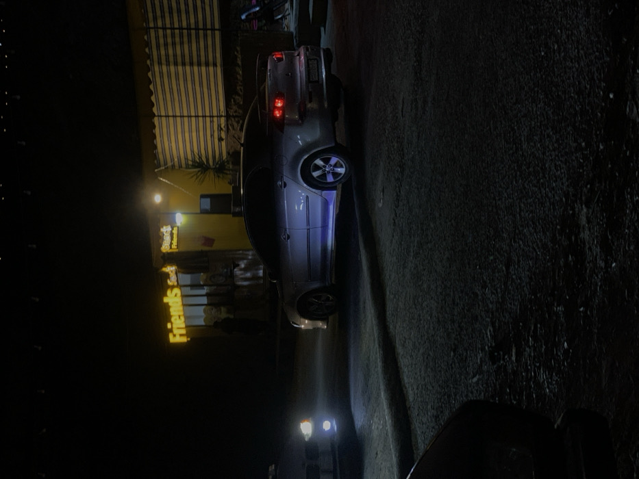 A silver car is parked at night near a building with an awning. The building has a sign that reads "Friends Cafe". Headlights from another vehicle illuminate the street in the foreground. The road surface is dark and textured. Overall, the image is dark with artificial lighting.