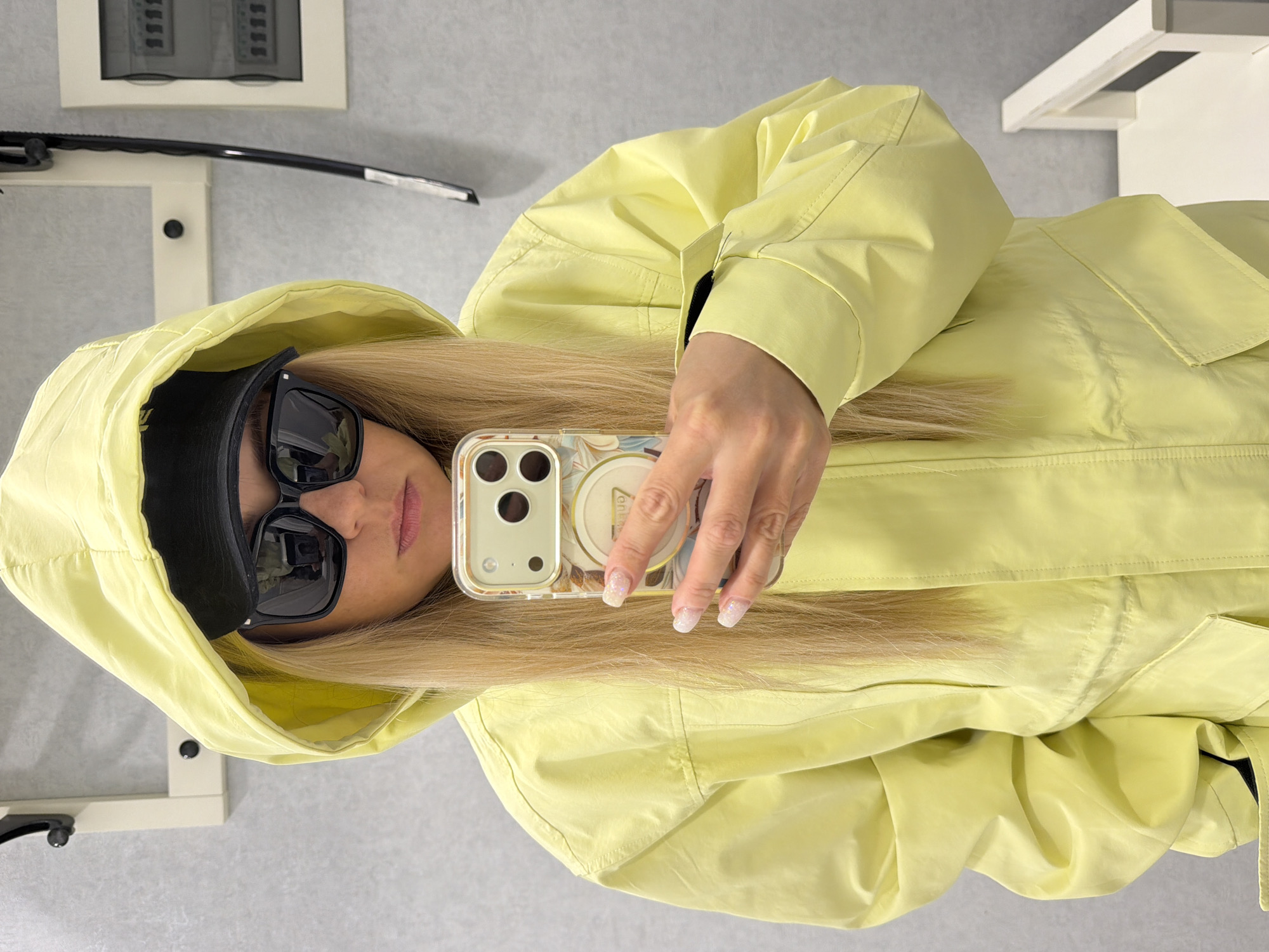 The image captures a mirror selfie taken by a person with long, straight blonde hair, located in Votkinsk, Russia. The person is wearing a bright, light yellow hooded jacket, with the hood up over a black baseball cap. They are also sporting large black sunglasses, which obscure their eyes. Their lips are visible, appearing to have a light pink color.
The individual is holding a smartphone, likely an iPhone given the camera setup, in their right hand. The phone is encased in a clear or light-colored protective case with a subtle, abstract, possibly floral or marbled pattern in beige and blue tones. A round pop socket or grip is attached to the back of the phone, featuring a design that includes some indistinct text, possibly "enamel" or similar, but it is partially obscured. The person's fingernails are manicured with a glittery or iridescent finish.
The setting appears to be indoors, against a plain, light-colored wall. In the background, to the upper left, an electrical panel or distribution board with visible switches is mounted on the wall. Below this, part of a white, possibly metallic, furniture piece or structure is visible. The overall scene suggests a functional or utility space rather than a decorative one. The lighting is even, suggesting indoor artificial light or diffuse daylight. The person's pose is casual, typical for a mirror selfie.