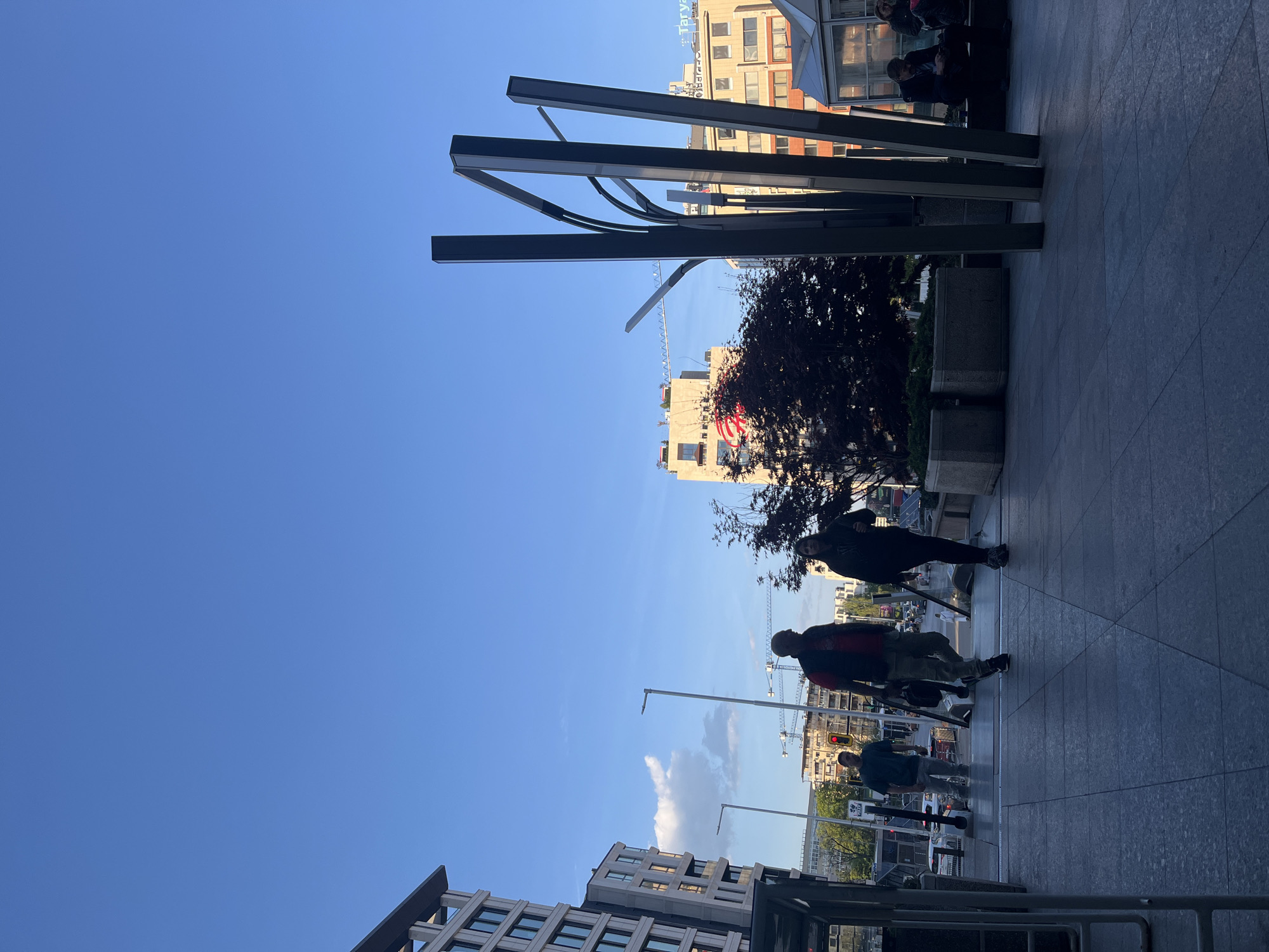 The image depicts a bustling urban street scene in Sofia, Bulgaria, captured during what appears to be the late afternoon or early evening on a clear, sunny day. The sky is a vibrant blue with a few scattered clouds, and the low sun casts long shadows across the paved surfaces.
The foreground features a wide, patterned gray pavement or plaza. Several individuals are visible, moving along the sidewalk. Two people are prominent in the midground, seen in partial silhouette: one wears a dark top and lighter pants, and the other a top with red and dark elements over lighter pants. Further back, near a building entrance, a small group of people is gathered, either sitting or standing.
The setting is characterized by modern architecture. Multi-story buildings with numerous windows line both sides of the street. On the left side, one building features a noticeable red circular logo or sign. On the right, another building facade clearly displays the text "Tarva BILD." Overhead, several striking, dark angular beams, possibly part of a contemporary shelter or artistic installation, extend across the frame.
In the background, tall construction cranes rise above the rooftops, indicating ongoing development in the city. Other urban elements include streetlights, concrete planters, and a visible red traffic light on the right. A dark-leafed tree adds a touch of nature to the otherwise concrete environment. The overall scene portrays a typical, dynamic city atmosphere.