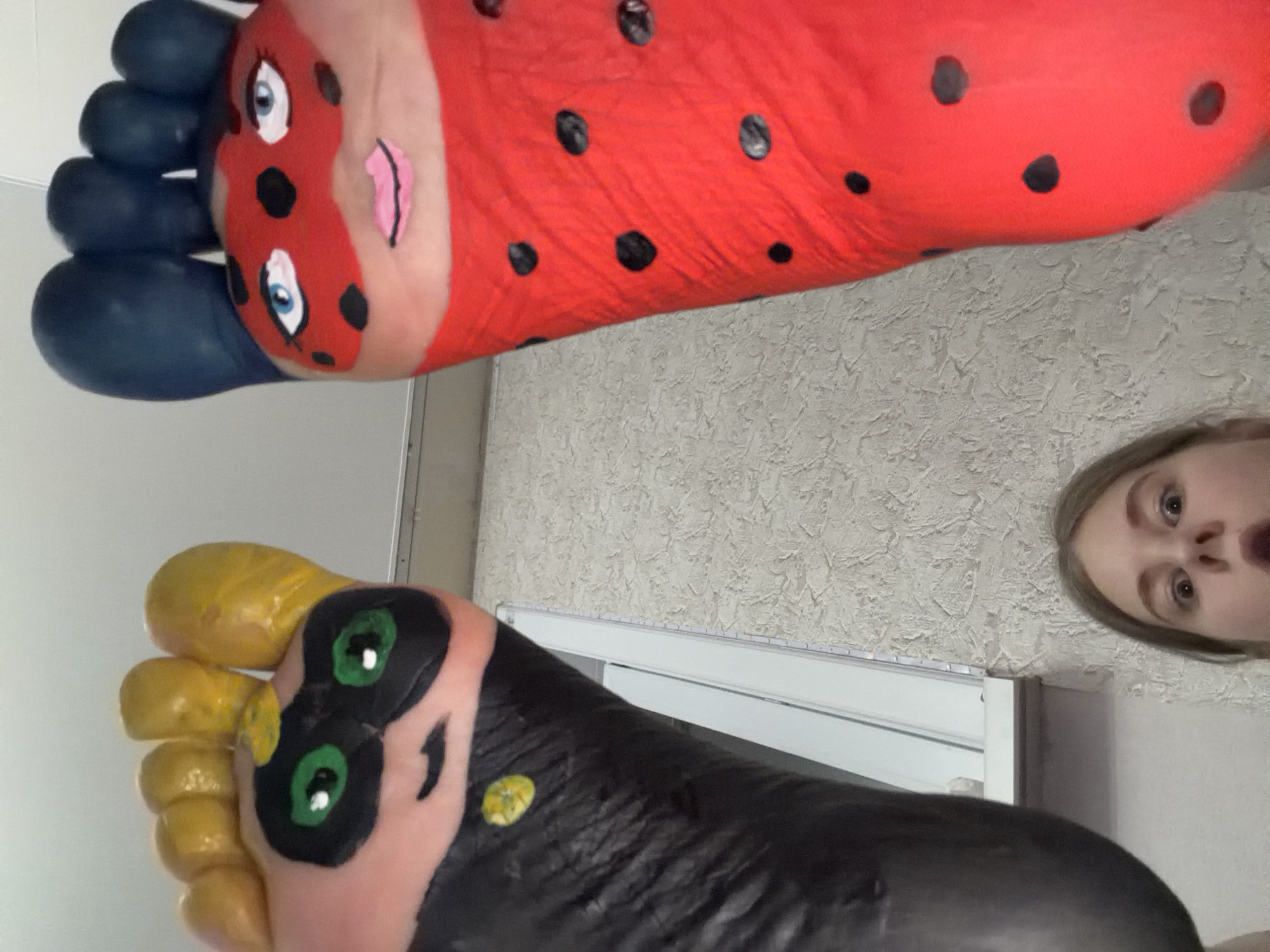 A low-angle shot shows two feet painted to resemble characters, with a person's face visible in the corner. The foot at the top is painted red with black spots and blue toes to resemble Ladybug from Miraculous Ladybug. The other foot is painted black with yellow toes and green eyes, likely representing another character from the same series. The background includes a patterned wall and a window frame. The image was likely taken indoors. No verifiable information is present to confirm this photo was taken in Perm, Russia.