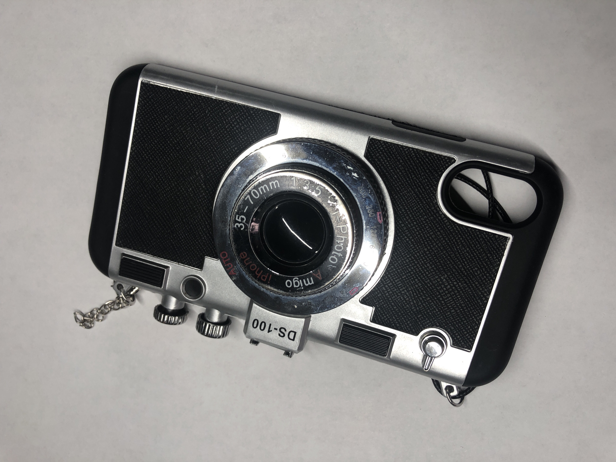 This media file showcases a silver and black iPhone case designed to resemble a vintage camera. The case is laid flat against a plain, light-colored surface, likely a table or backdrop, with no discernible background details to indicate a specific location. The object is oriented diagonally.
The iPhone case features a central lens-like embellishment with detailed markings, including "35-70mm," "f/3.5," and the text "iPhone" and "Amigo Photo" printed around it. Various dials and buttons are also visible on the case's surface, mimicking the controls of a traditional camera. These include two silver rotary knobs at the bottom left and a smaller silver button with a loop at the bottom right. A small, silver chain is attached to the left side of the case, and another loop for attachment is visible on the right.
Inscribed on a silver rectangular plate at the bottom center of the case is the text "DS-100" in black, stylized font, appearing upside down in the image. The textured black sections on the case likely represent the grip of the camera. The overall aesthetic suggests a playful and retro design for a smartphone accessory. There is no indication of people, weather, or time of day, and the setting is neutral.