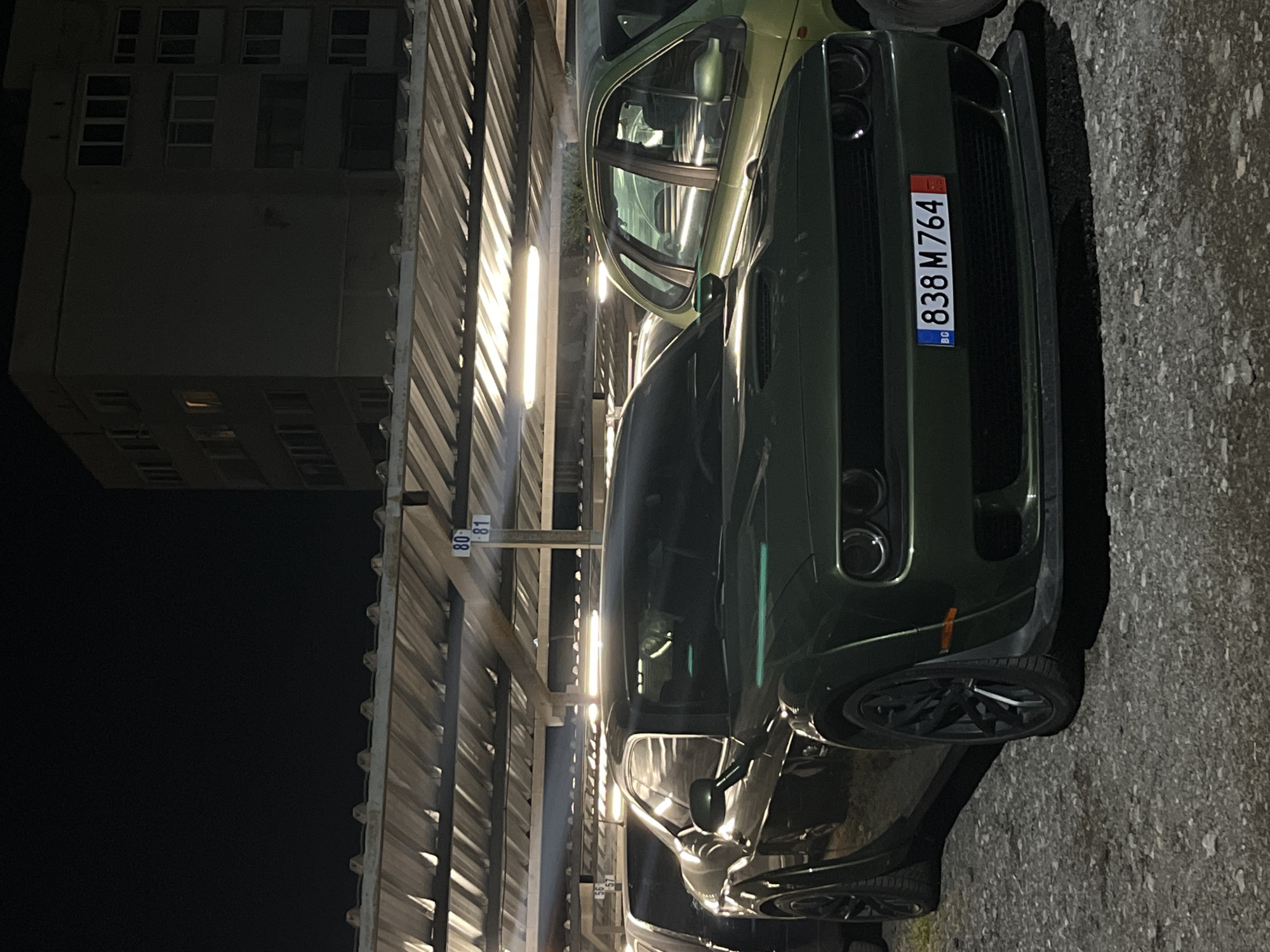 The image shows the front of a dark green Dodge Challenger parked in what appears to be an underground parking garage. The car has a Bulgarian license plate that reads "838 M764" and the expiration date "22" on it. The license plate indicates that the car is registered in Sofia, Bulgaria. The background includes the structure of the parking garage ceiling, lighting fixtures, and a building illuminated in the dark. The focus of the picture is on the car's front end, highlighting its design.