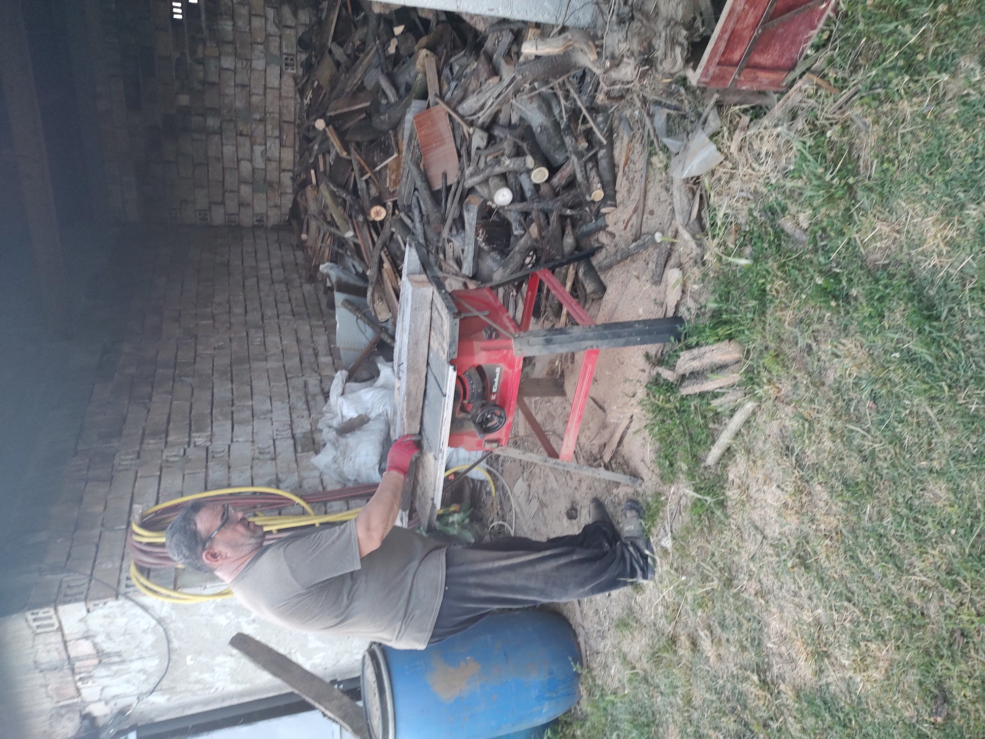 A man wearing glasses, a grey t-shirt, dark pants, and red gloves is standing in front of a red table saw. He is holding a piece of wood and appears to be working with the saw. The saw is set up outdoors in what looks like a backyard or workshop area. Behind the man and the saw is a large pile of logs and wood debris, as well as a blue plastic barrel. The background shows a brick wall and some industrial piping. The overall scene suggests a DIY or woodworking activity taking place in an outdoor, somewhat cluttered environment. This location is identified as being in the city of Kochmar, Bulgaria.