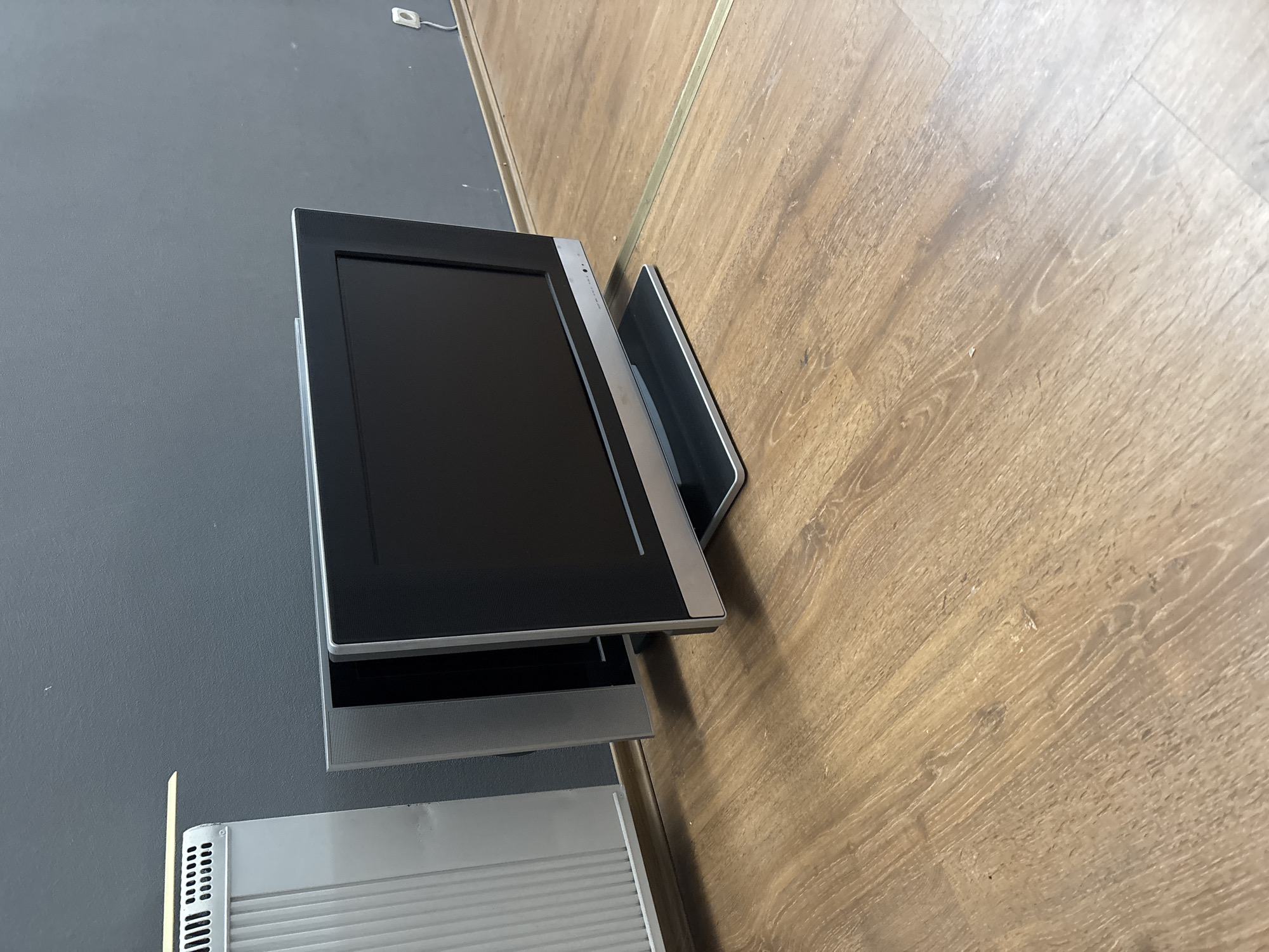 A flat-screen television is positioned on a wooden floor in front of a dark grey wall. To the left of the television, a silver radiator is visible. The television is black with a silver trim and stand. A thin golden strip runs across the floor from the base of the television towards the right side of the image. The overall setting appears to be an indoor space, possibly a room within a building in Zaporizhzhia, Ukraine. There are no people or animals present in the image. The lighting suggests an artificial light source, casting subtle shadows on the floor.
