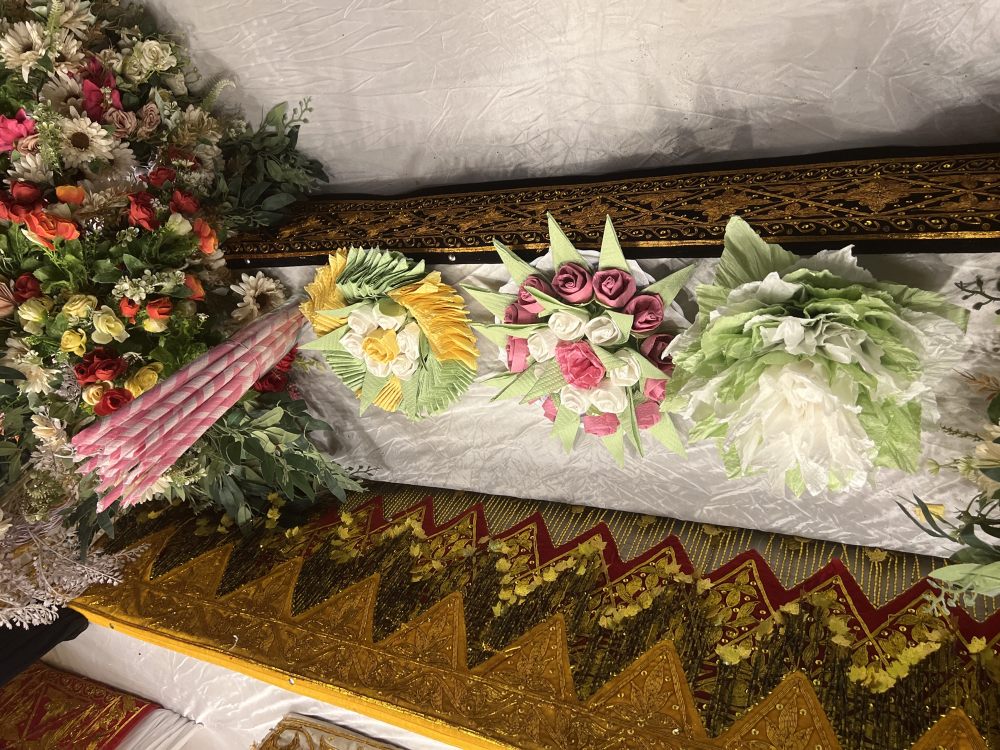 This image depicts a close-up, overhead view of an ornate, likely ceremonial, arrangement. Dominating the composition is a large floral arrangement featuring a mix of red, orange, yellow, pink, and white artificial flowers interspersed with green foliage. To the left of the floral arrangement, a piece of fabric with a striking gold and red geometric pattern is visible, suggesting a rich and possibly traditional textile. Further down the arrangement, there are three smaller, clustered bouquets of artificial flowers. The bottom bouquet is predominantly white and green, with the other two featuring pink and white roses, and yellow and white flowers respectively. The entire scene is set against a textured, off-white fabric background. The lighting appears to be artificial, casting subtle shadows. No people are visible in the image, and no specific location within Baratpayaitek, Indonesia, can be definitively identified from the visual cues alone. The scene suggests a decorative setup, possibly for an event or a display.