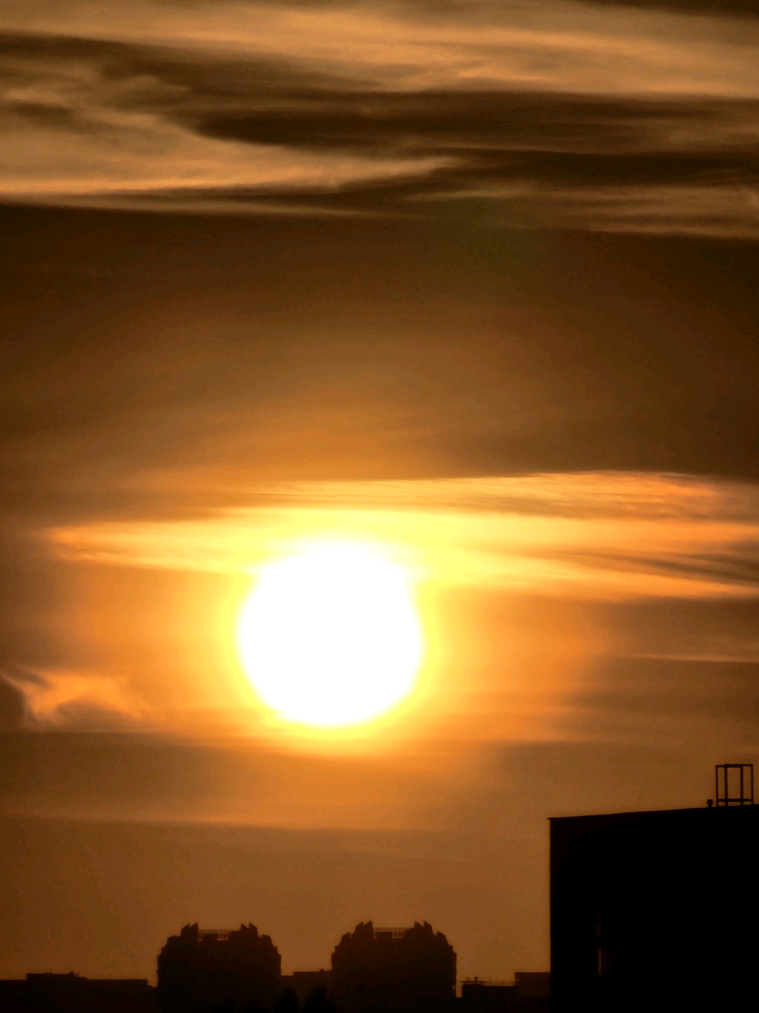 An extreme close-up shot captures a bright, luminous sun positioned in the upper center of the frame, casting a warm, orange glow across the sky. Wispy, horizontal clouds are visible, illuminated by the sun's intense light. In the foreground, the dark silhouettes of buildings stand out against the vibrant sky, suggesting an urban setting. The building on the right is taller and features a distinct, skeletal structure on its roof. Two smaller, silhouetted structures are visible on the left side of the frame. The overall impression is a dramatic sunset or sunrise over the city of Khimki, Russia.