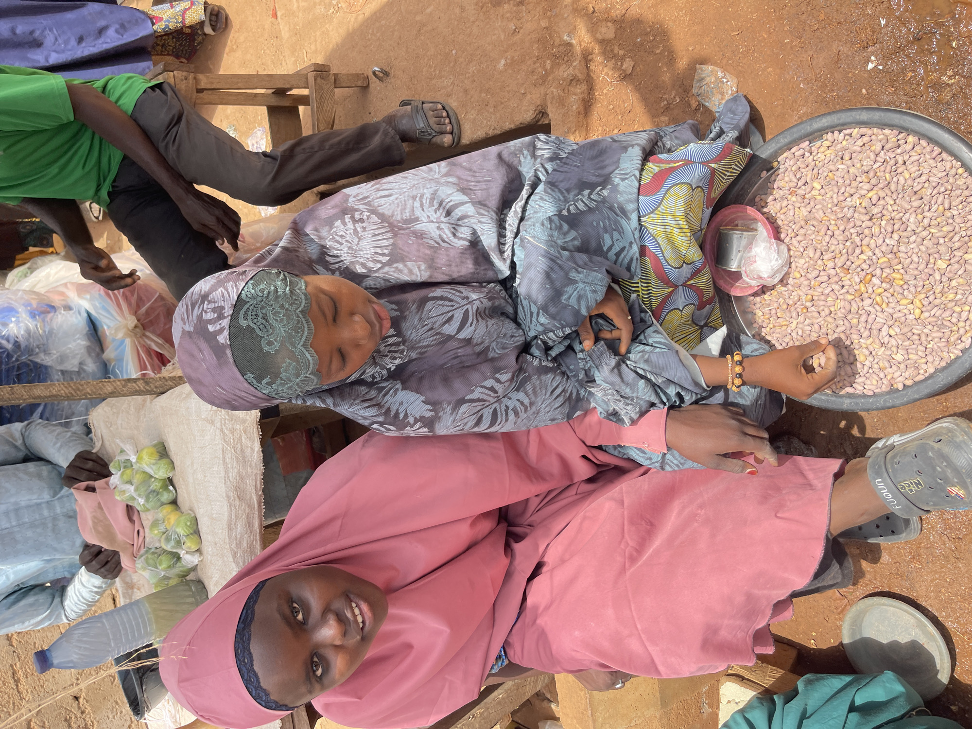 This image, taken in Zaria, Nigeria, depicts an outdoor market scene under bright daylight. Three women and one man are present, engaged in commercial activity.
In the foreground, a woman wearing a pink hijab and matching long-sleeved top smiles warmly directly at the camera. She appears to be sitting on the ground. Next to her, slightly behind, is another woman dressed in a grey-blue patterned long dress and a matching hijab with green lace detailing. Her eyes are closed or looking down, and her right hand is near a large black basin overflowing with numerous peanuts. A small red plastic container holding a metal cup sits within the basin of peanuts.
To the left and further back, a third woman, partially visible in a blue-grey garment, attends to small clear plastic bags filled with green limes or small lemons, arranged on a white woven mat. Adjacent to this area, a man wearing a bright green t-shirt and dark trousers is seated on a wooden stool, with only his legs and arms clearly visible.
The ground is a light brown, sandy or earthen surface, indicating an outdoor setting. Other objects visible include a plastic water bottle and a grey metal plate on the ground. The attire of the individuals, particularly the hijabs and light clothing, is consistent with a warm climate. On a grey croc-style sandal worn by one person, the word "Funun" is distinctly visible. The scene portrays a typical bustling local market environment.