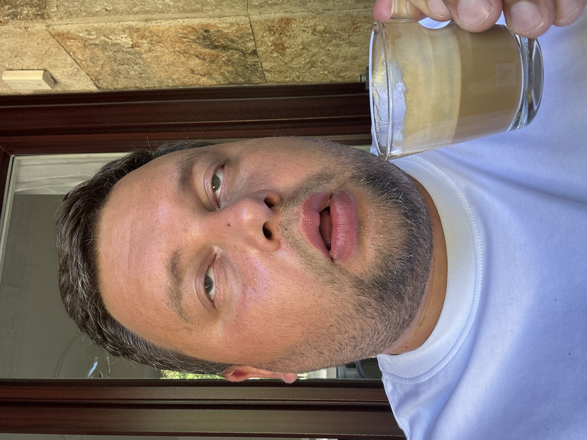 A man with fair skin, short dark hair, and a stubbled beard is holding a glass of layered coffee. He is wearing a white shirt and looking directly at the camera with slightly open mouth. Behind him is a stone wall, a dark brown wood frame. A window can be seen. The city or specific location in Sant Feliu de Guíxols, Spain, cannot be determined from the image alone.