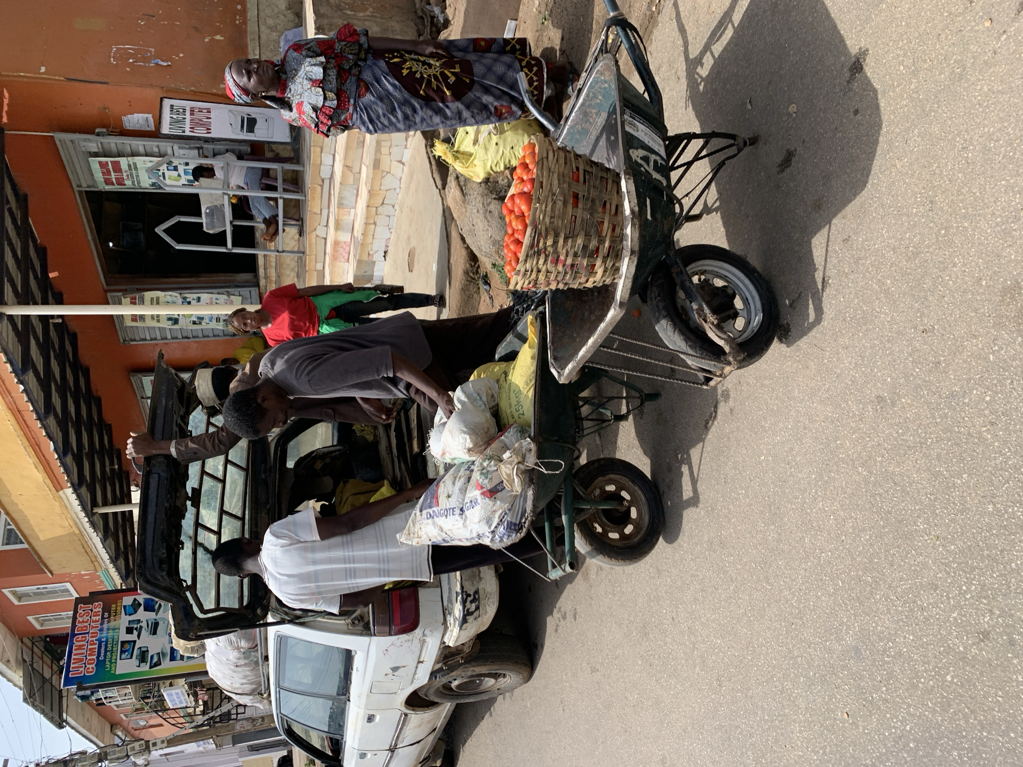 The image shows a bustling scene in Jos, Nigeria. A white car with its trunk open is being loaded with goods. A man in a gray shirt and a man in a white shirt are actively loading bags labeled "DAIGOTE GAR" into the car. A wheelbarrow filled with tomatoes in a woven basket, along with other goods, is also part of the load. An older woman in a patterned dress and headwrap stands beside the wheelbarrow. Several other people are present in the background, including a person in a red shirt and another in a green shirt. A sign for "LIVING BEST COMPUTERS" is visible above the car, indicating a local business. The environment appears to be a street or marketplace.