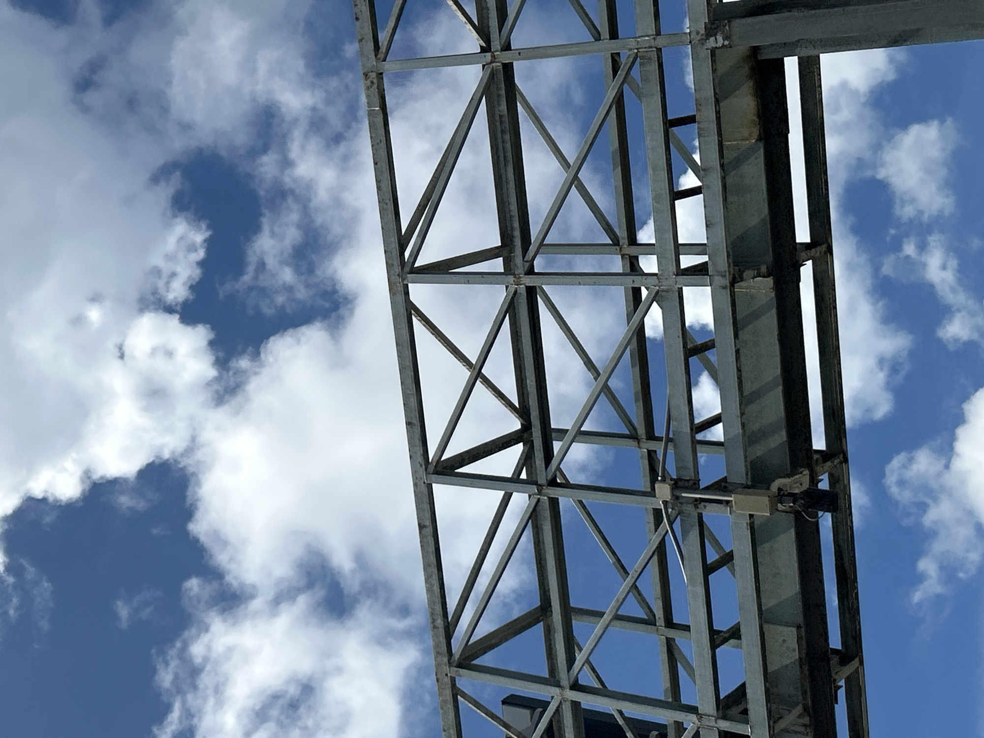 A close-up, upward-facing shot captures the underside of a large, metallic overhead sign structure against a vibrant blue sky filled with scattered white clouds. The structure is a complex lattice of steel beams, forming a series of triangular and rectangular supports. The metal appears weathered and industrial, with visible signs of rust and age. Attached to the underside of the structure are various pieces of equipment, including what appear to be cameras or sensors, mounted on brackets and wires. The sky dominates the background, with the clouds creating dynamic patterns and shadows across the scene. The location in Wichit, Thailand, is not explicitly discernible from the visual elements of the image itself.