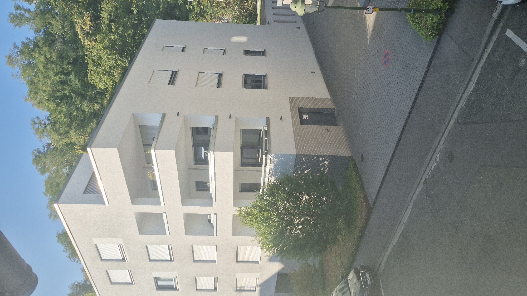 A modern, multi-story apartment building is seen from a high angle, partially obscured by a grey pipe or overhang at the top of the frame. The building features a white facade, numerous balconies with glass railings, and windows with closed white shutters. A small tree with green foliage is situated in front of the building, casting a shadow. The building is positioned on a slight incline, with a paved area in front of it, possibly a parking lot or walkway, marked with a red circular symbol. A dark-colored Audi car is parked to the left, with its license plate visible. Behind the building, a dense, green forest covers a hillside under a clear blue sky. A street runs in front of the property, with a curb and drainage grate visible. A lamppost with a green fixture stands near the right edge of the frame, next to a bush and a sign indicating parking restrictions. The overall scene depicts a residential area in Baden-Baden, Germany, on a bright, sunny day.
