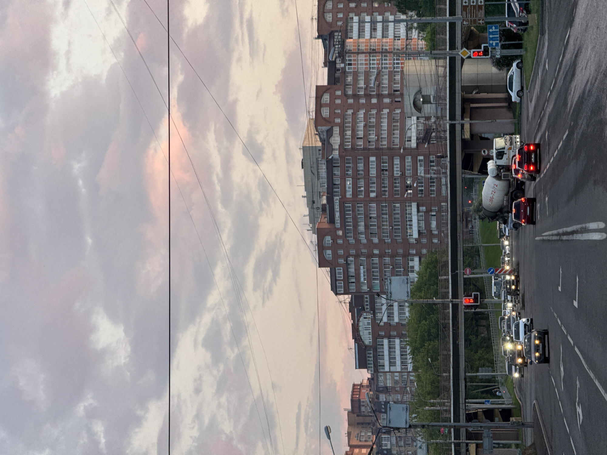 The image depicts an urban scene in Krasnoyarsk, Russia, during what appears to be twilight or early evening. A multi-lane road occupies the foreground, with several cars moving in various directions. A cement truck is prominently visible on the right side of the road. Overhead, a road overpass is supported by concrete pillars, with trees and greenery visible beneath it. In the background, a series of multi-story brick apartment buildings with numerous windows line the horizon. The sky above is filled with soft, pastel-colored clouds, tinged with pink and orange hues, suggesting either sunrise or sunset. Power lines crisscross the sky. There are no individuals clearly visible in the image.