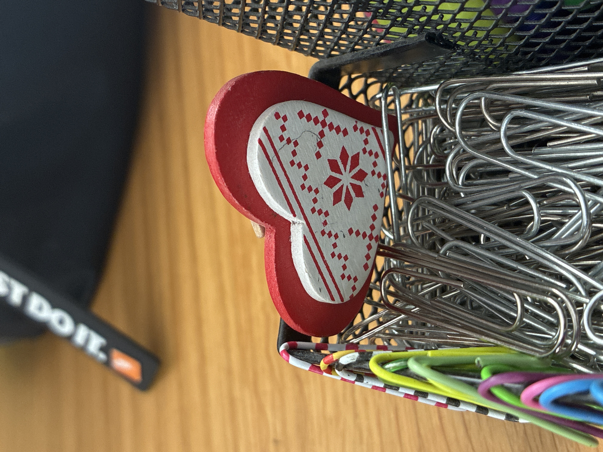 A close-up shot shows a red and white heart-shaped paperclip on a desk. The heart is adorned with a snowflake pattern in red. The paperclip is placed in a black mesh container filled with silver paperclips. In the background, out of focus, is a black object with the words "JUST DO IT." printed in white. The desk surface is made of wood. The image is captured from a slightly elevated angle.