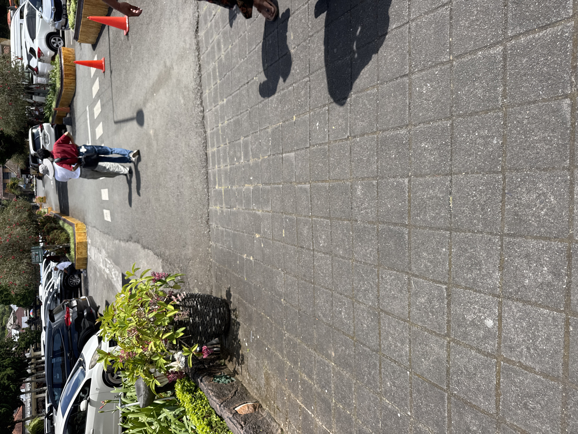 The image shows a street scene in Baturiti, Indonesia. The street is paved with gray square stones. A potted plant sits to the left side of the frame near the curb. Several cars are parked along the curb, varying in color, including white, black, and blue. In the background, two individuals wearing a red shirt and a white shirt are seen walking on the street. Traffic cones are placed to the left and in the background. A person's feet and shadow are visible at the top of the frame.